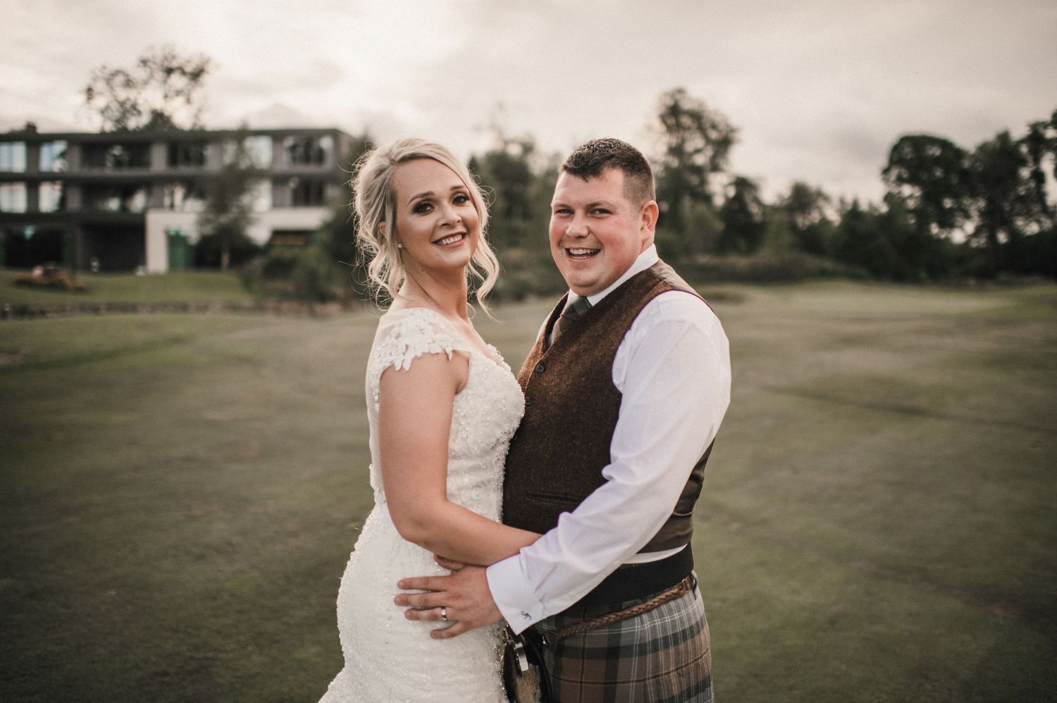 Newlyweds pose together on golf course, groom wearing traditional Scottish kilt.