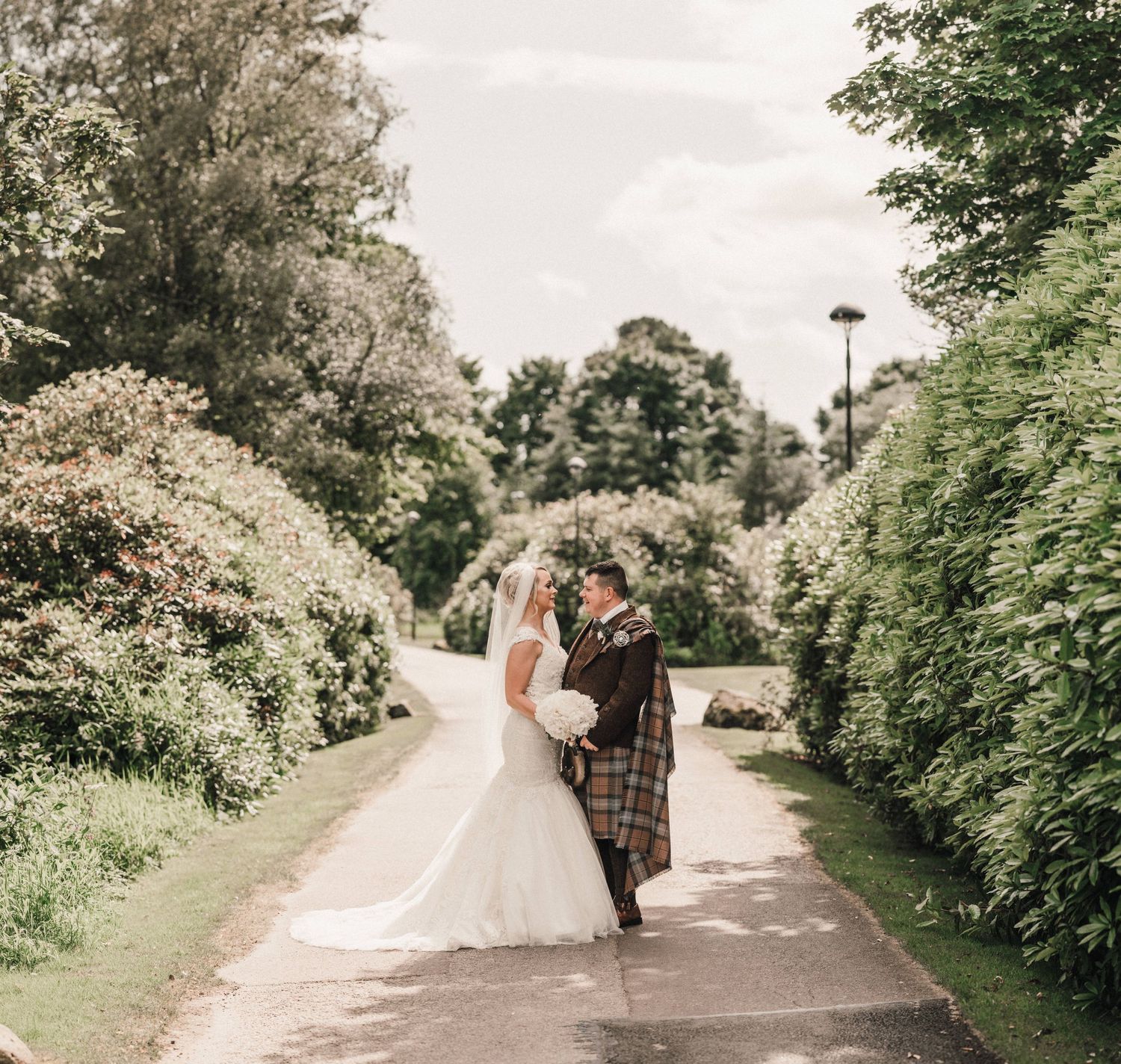 A couple in wedding attire walks along a tree-lined garden path on their special day.