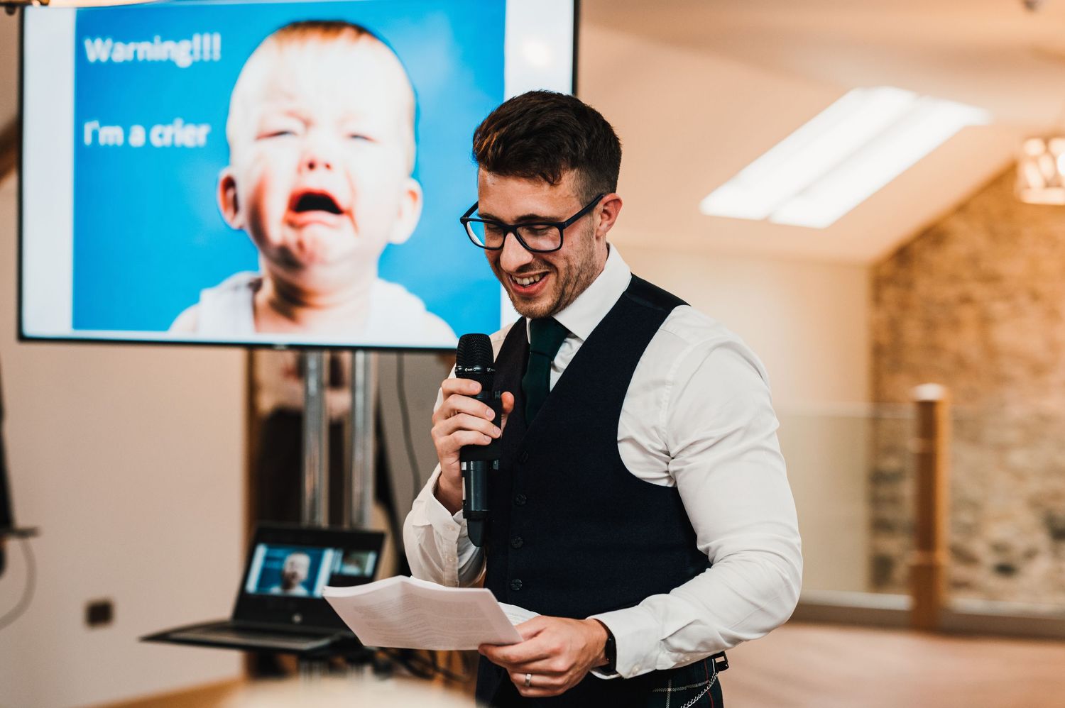 Speaker reads from notes during wedding reception with presentation screen in background.