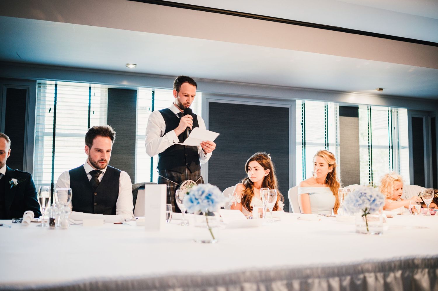 A wedding reception scene with guests seated at a long table while someone delivers a speech.