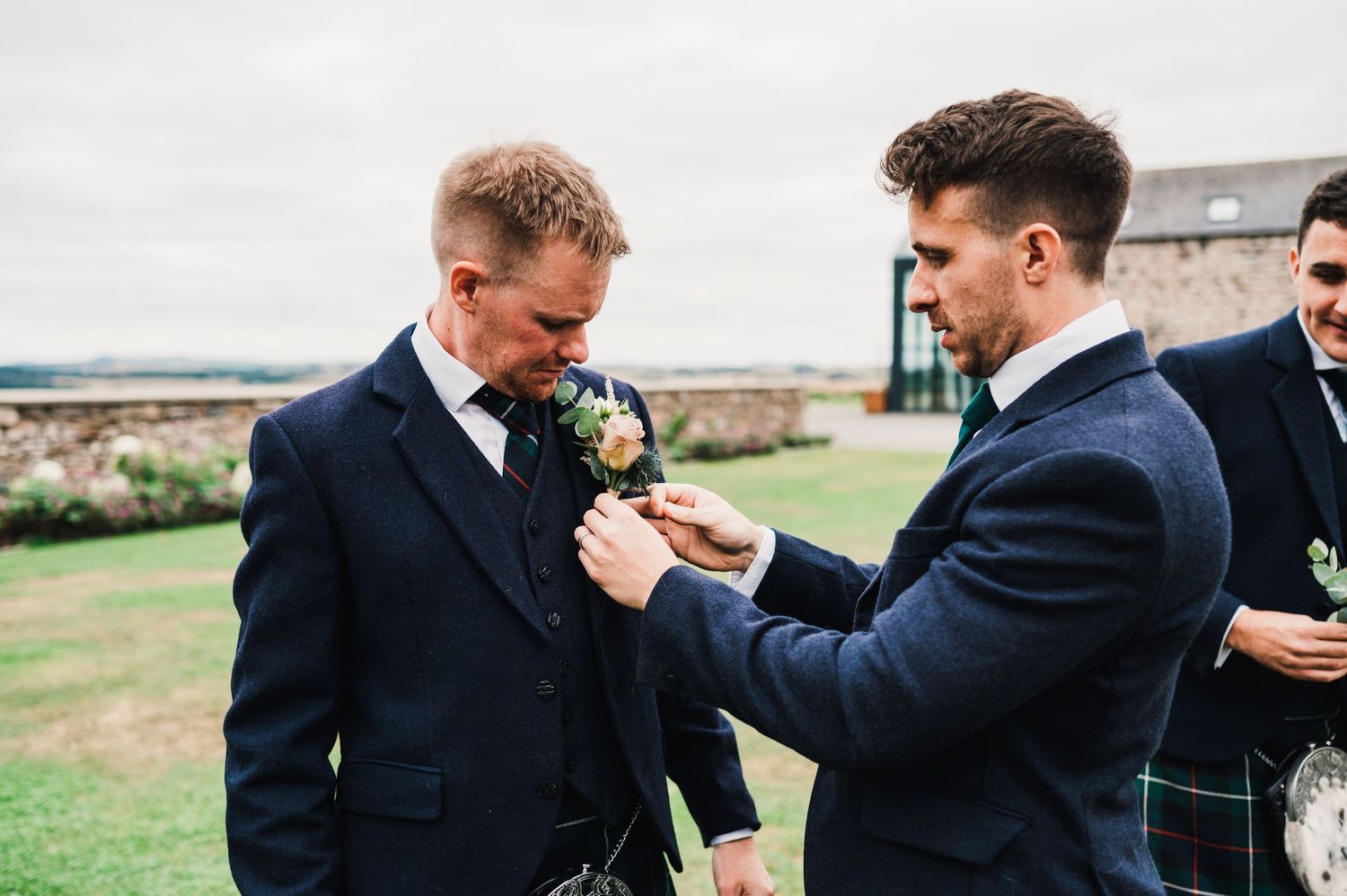 Two people in formal suits pin boutonnieres outside at a wedding venue.
