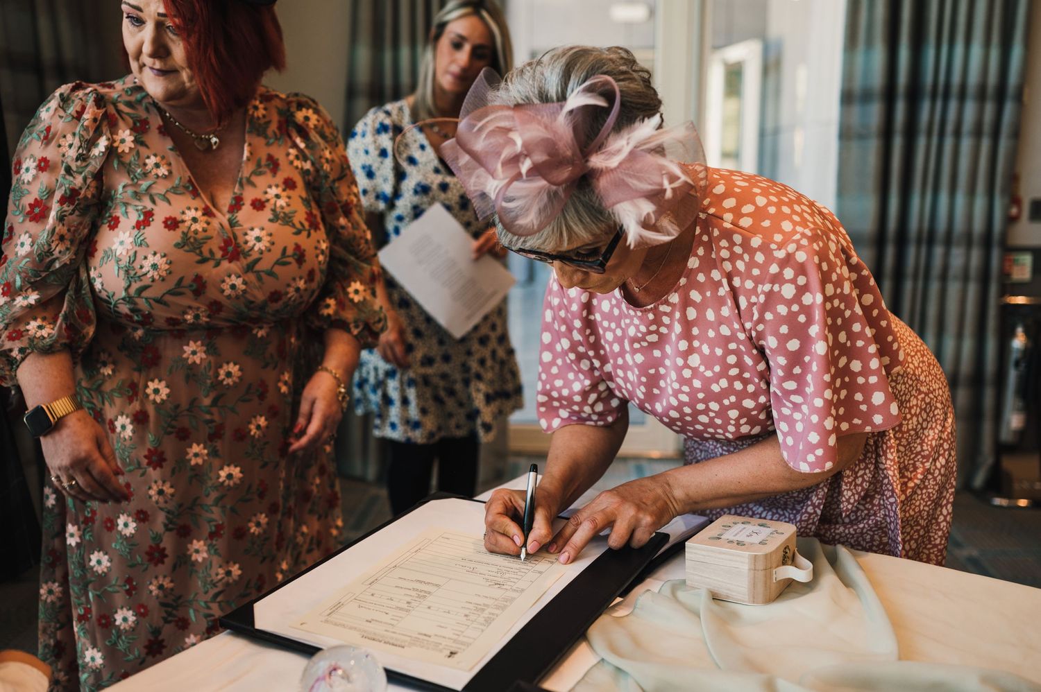 Wedding guests in colorful floral dresses signing formal documents at an indoor venue.