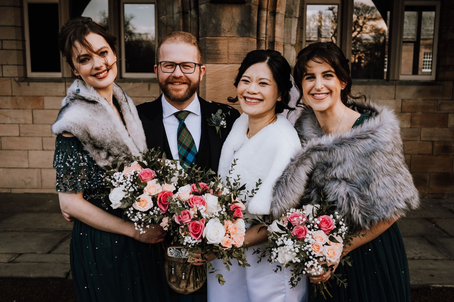 A wedding party of four wearing fur shawls and formal attire pose with pink floral bouquets.
