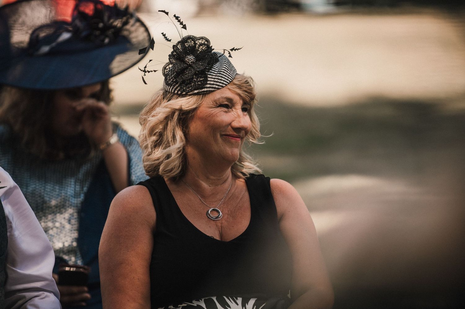 Wedding guest in formal black dress and ornate fascinator smiles during celebration.