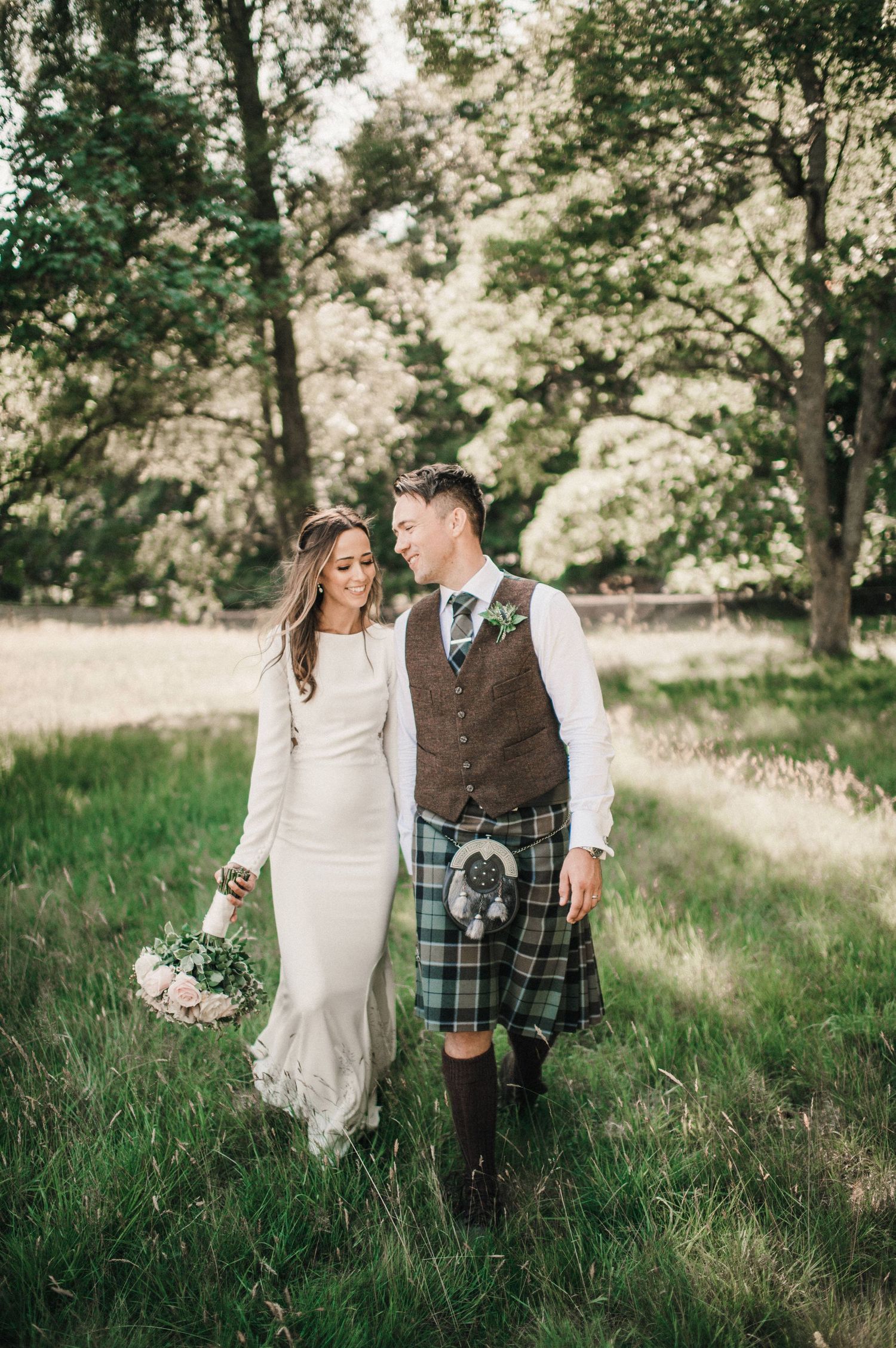 Newlyweds walk hand in hand through a lush green forest, with the groom in traditional Scottish highland dress.