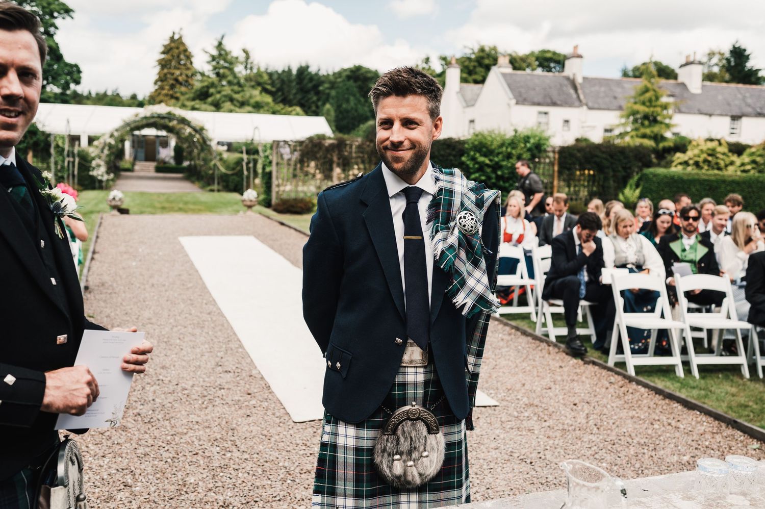 Attendee in traditional Scottish kilt stands at outdoor wedding ceremony with white chairs.