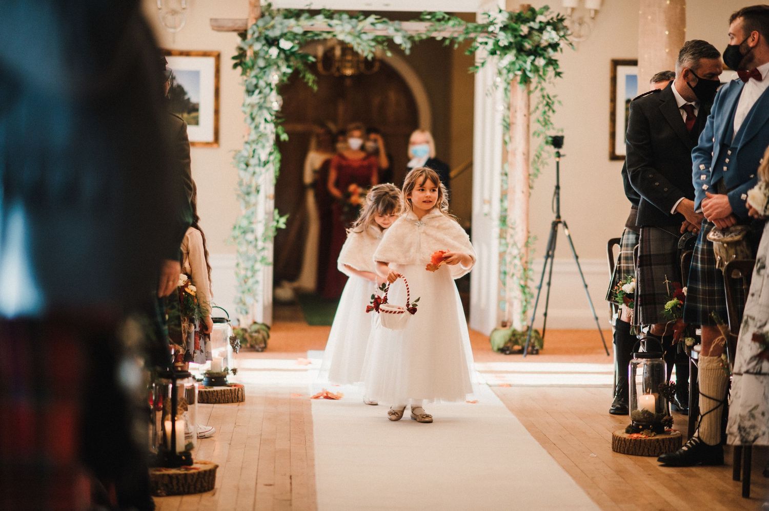 Young flower girls in white dresses walk down the aisle under an elegant floral archway.