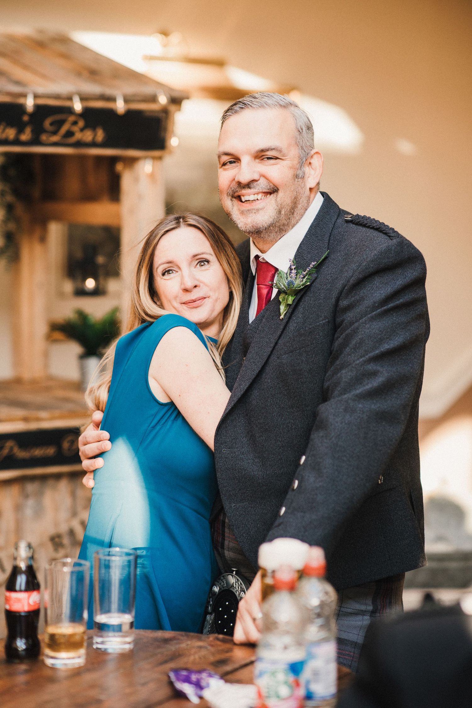 A couple embraces at an indoor venue, with the person in a blue dress leaning against their partner in a black suit.