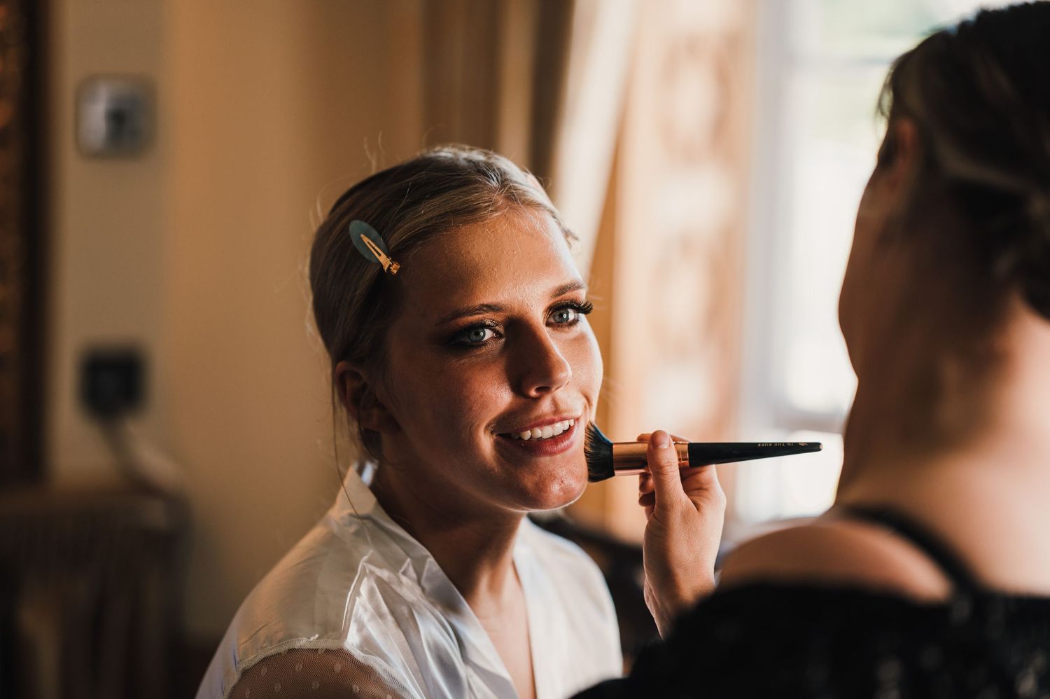 Makeup application during bridal preparation with natural window lighting.