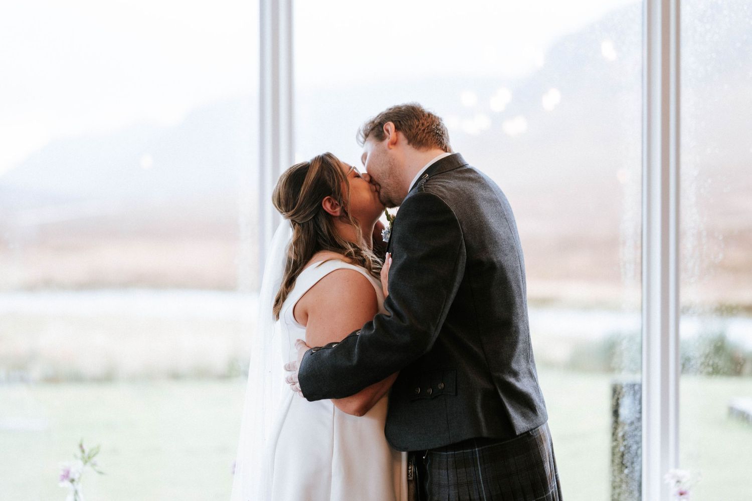 Romantic couple embraces and kisses in front of large windows with scenic mountain landscape in background.