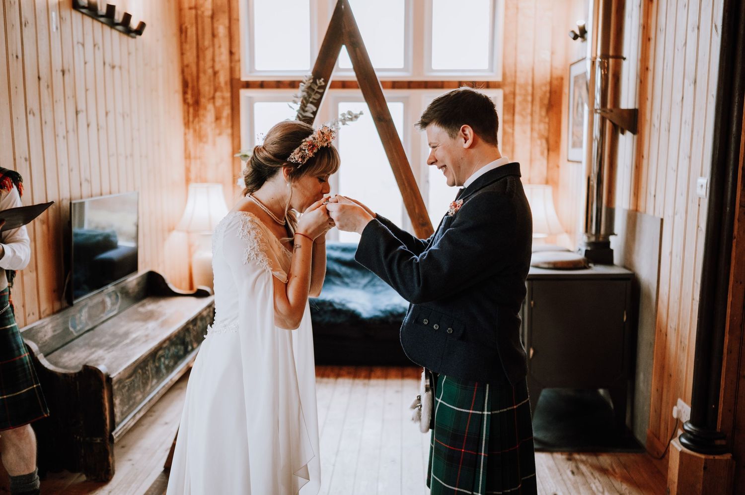 A couple shares an emotional moment in traditional Scottish wedding attire by a window.
