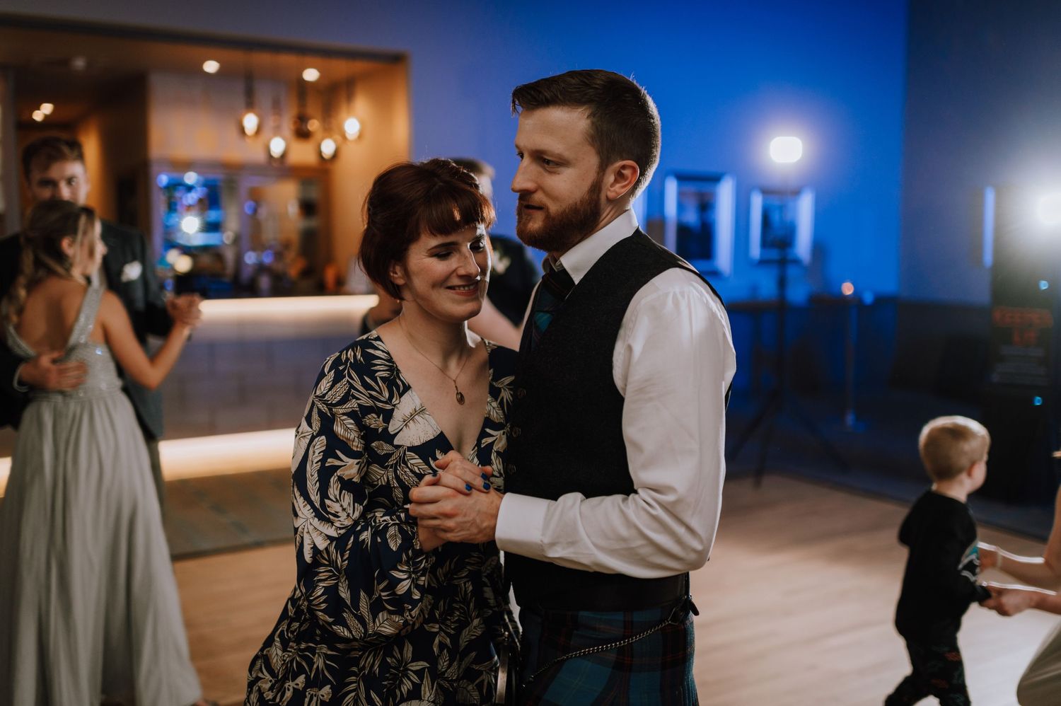 Couples slow dance together on a reception floor with blue ambient lighting.