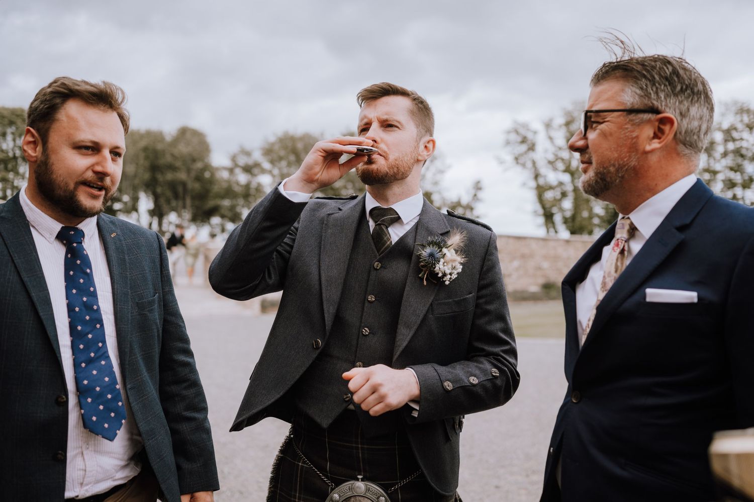 Wedding guests in formal attire share drinks and laughter outdoors.