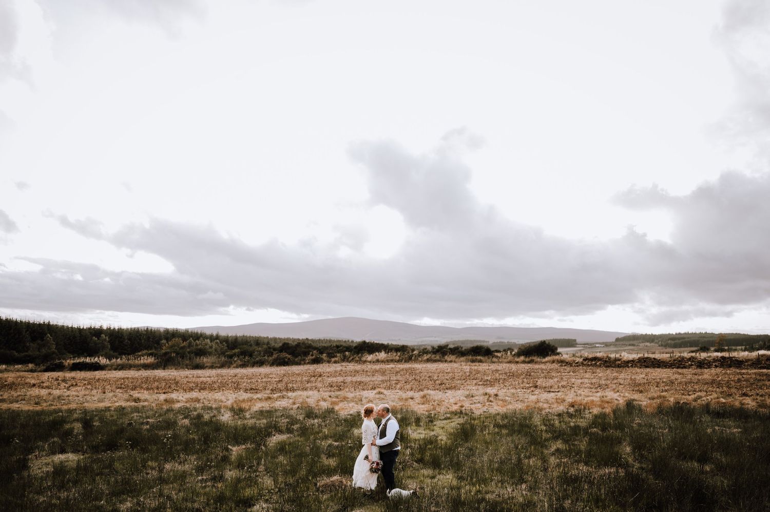 A couple stands together in a vast open field with dramatic cloudy skies and rolling hills.