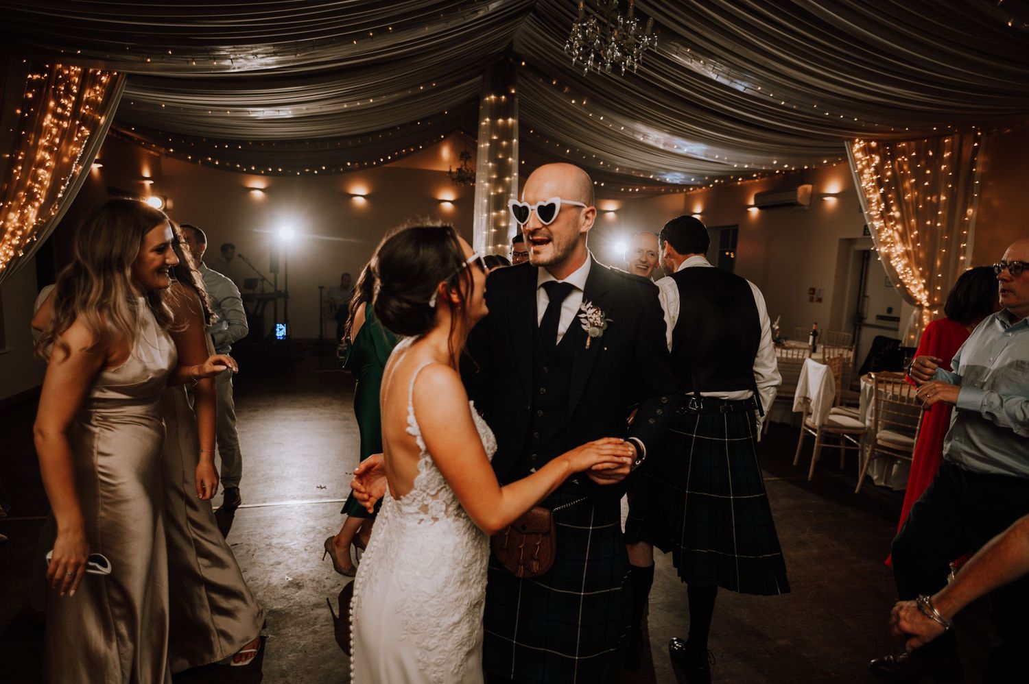 Wedding guests dance together on a dimly lit dance floor decorated with hanging lights and fabric draping.