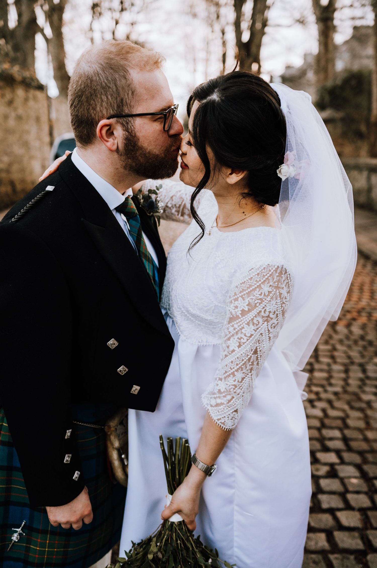 A romantic wedding moment with the groom in a traditional kilt and the bride in a lace-sleeved white dress on a cobblestone path.
