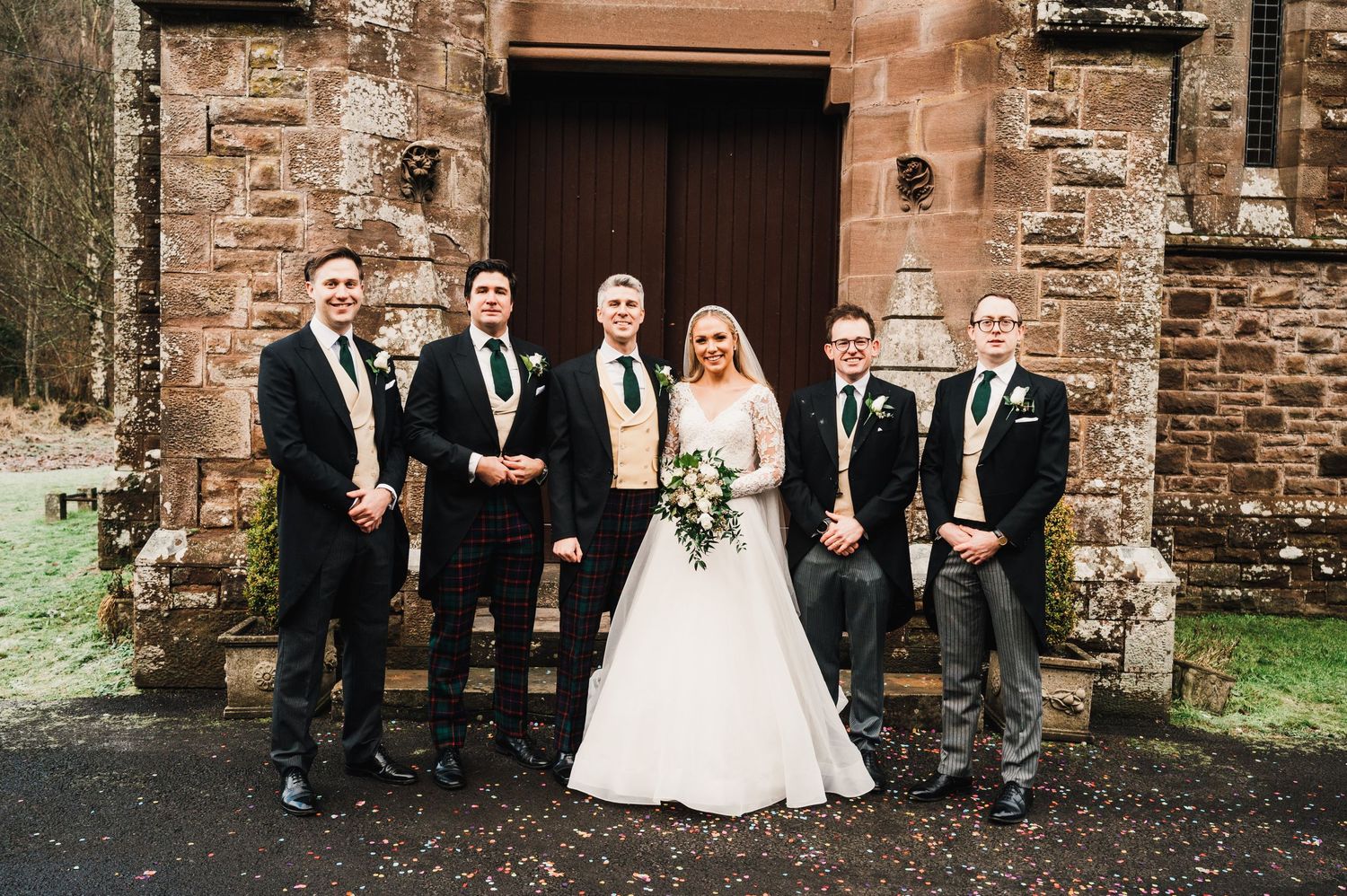 Wedding party in black suits poses outside historic stone church entrance.