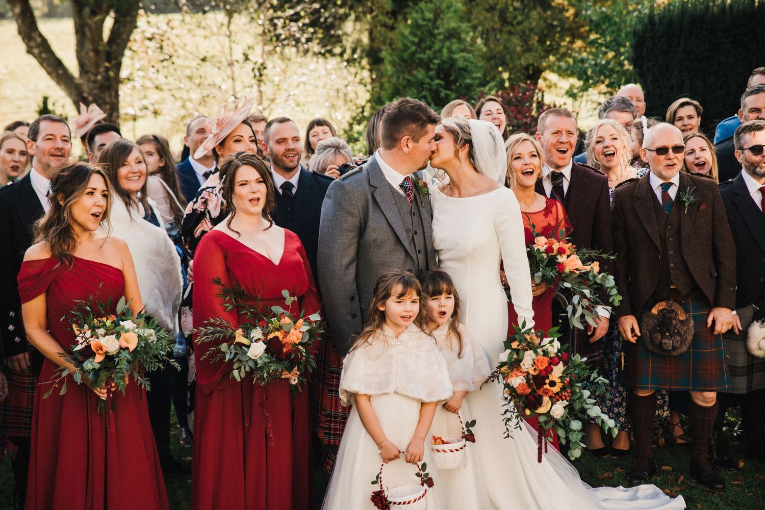 Outdoor wedding ceremony with bridal party in burgundy dresses and traditional Scottish attire.