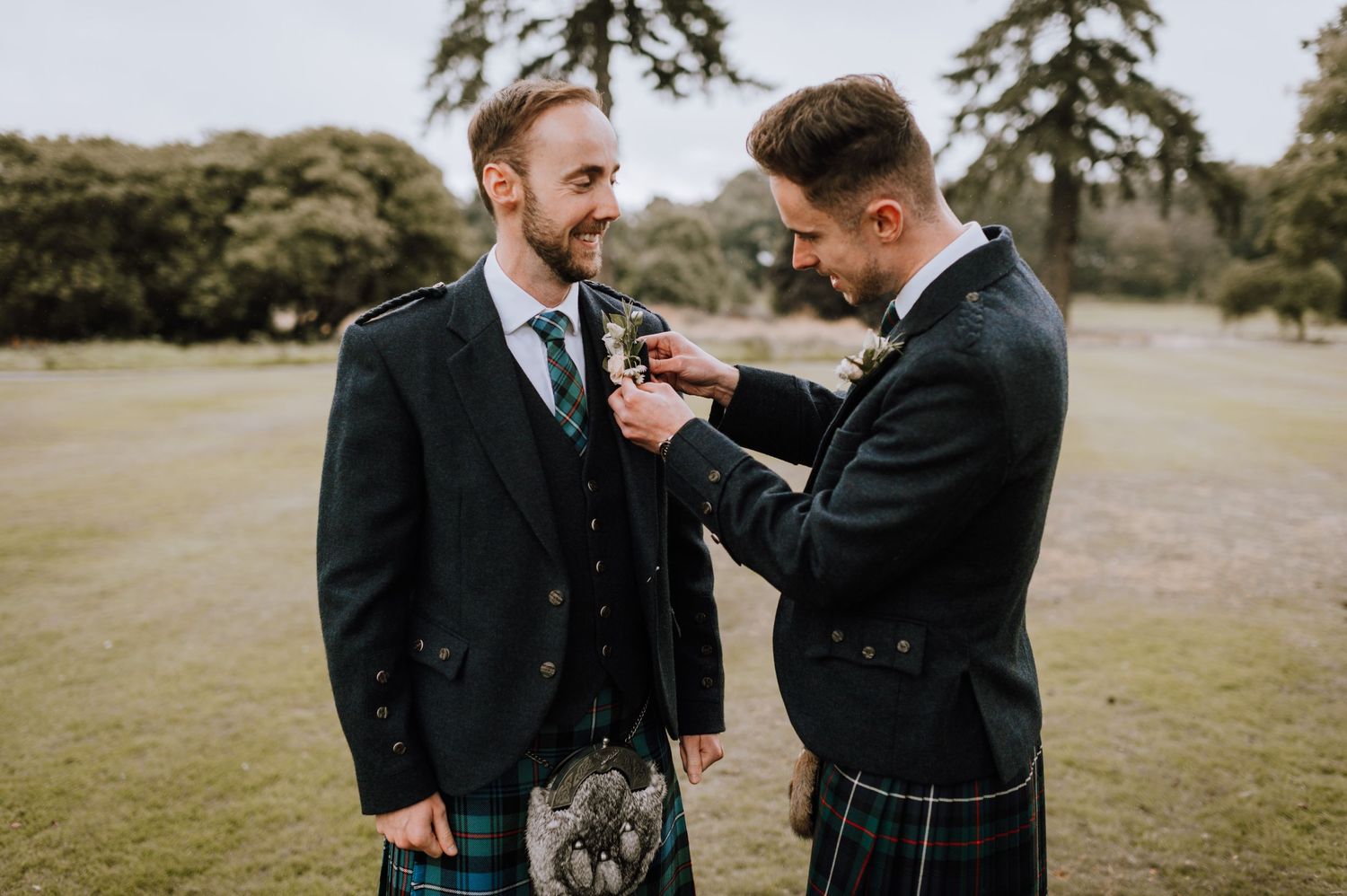 Men in traditional Scottish formal wear helping with a boutonniere outdoors.