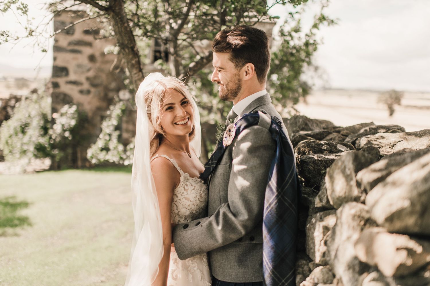 Sunlit wedding portrait against a stone wall with natural landscape in background.