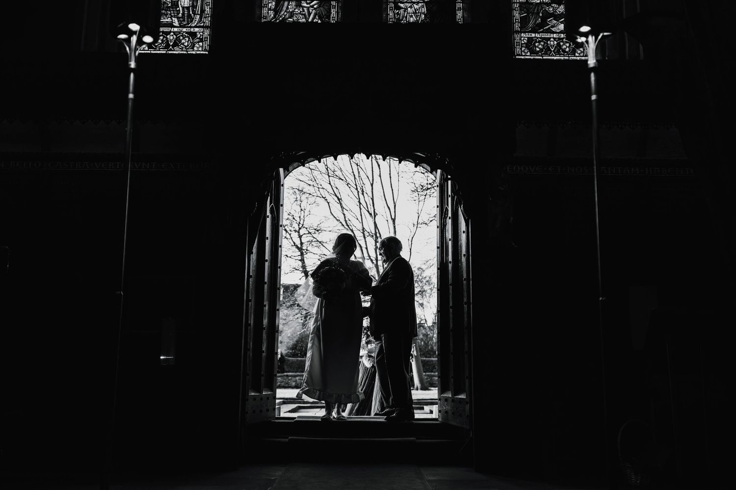 Silhouetted figures stand in a gothic church doorway creating dramatic wedding ceremony composition.
