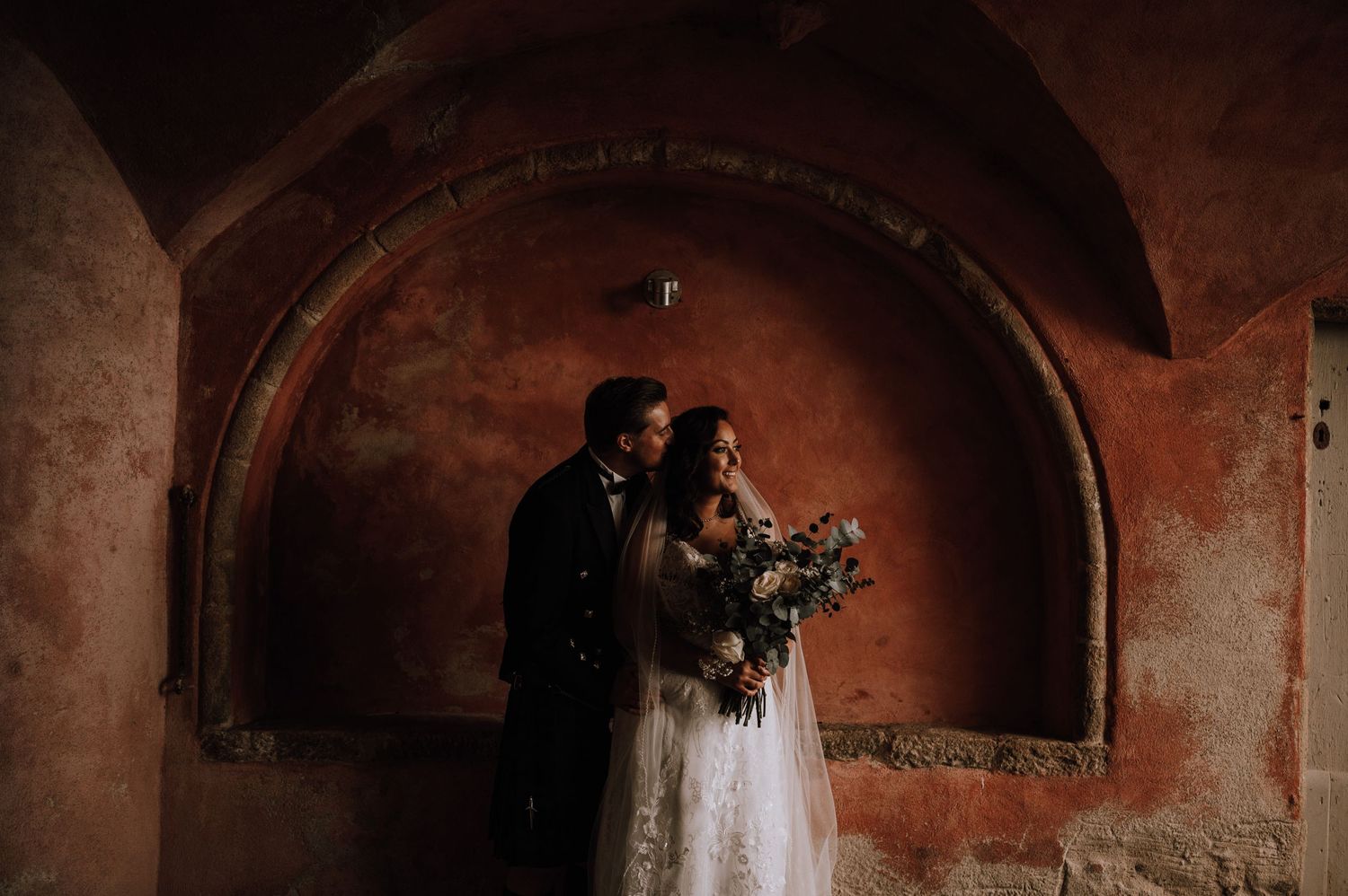 A romantic wedding portrait under an arched stone alcove with moody lighting.