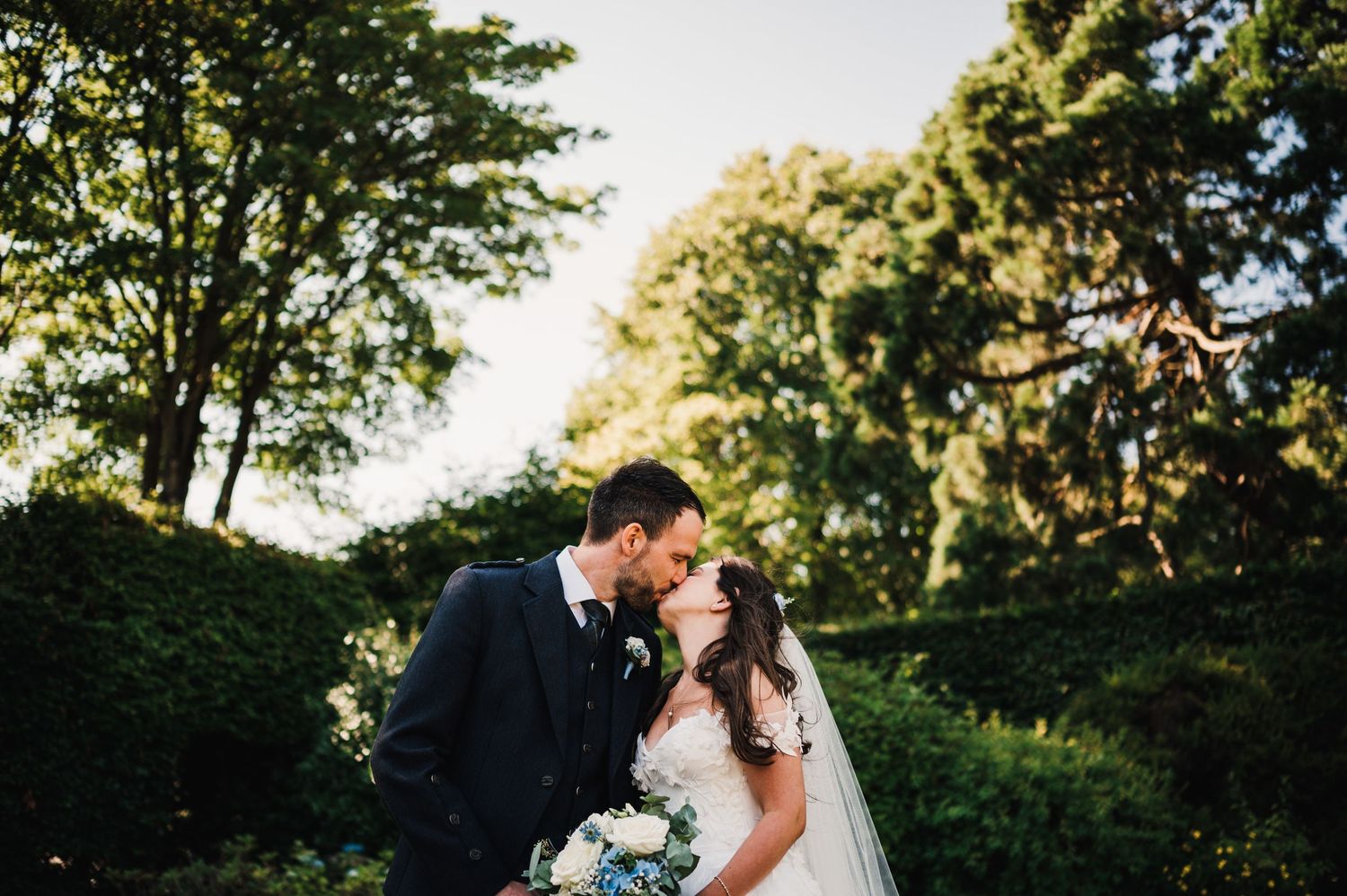 A romantic wedding portrait of newlyweds embracing in a lush garden setting with soft sunlight filtering through trees.