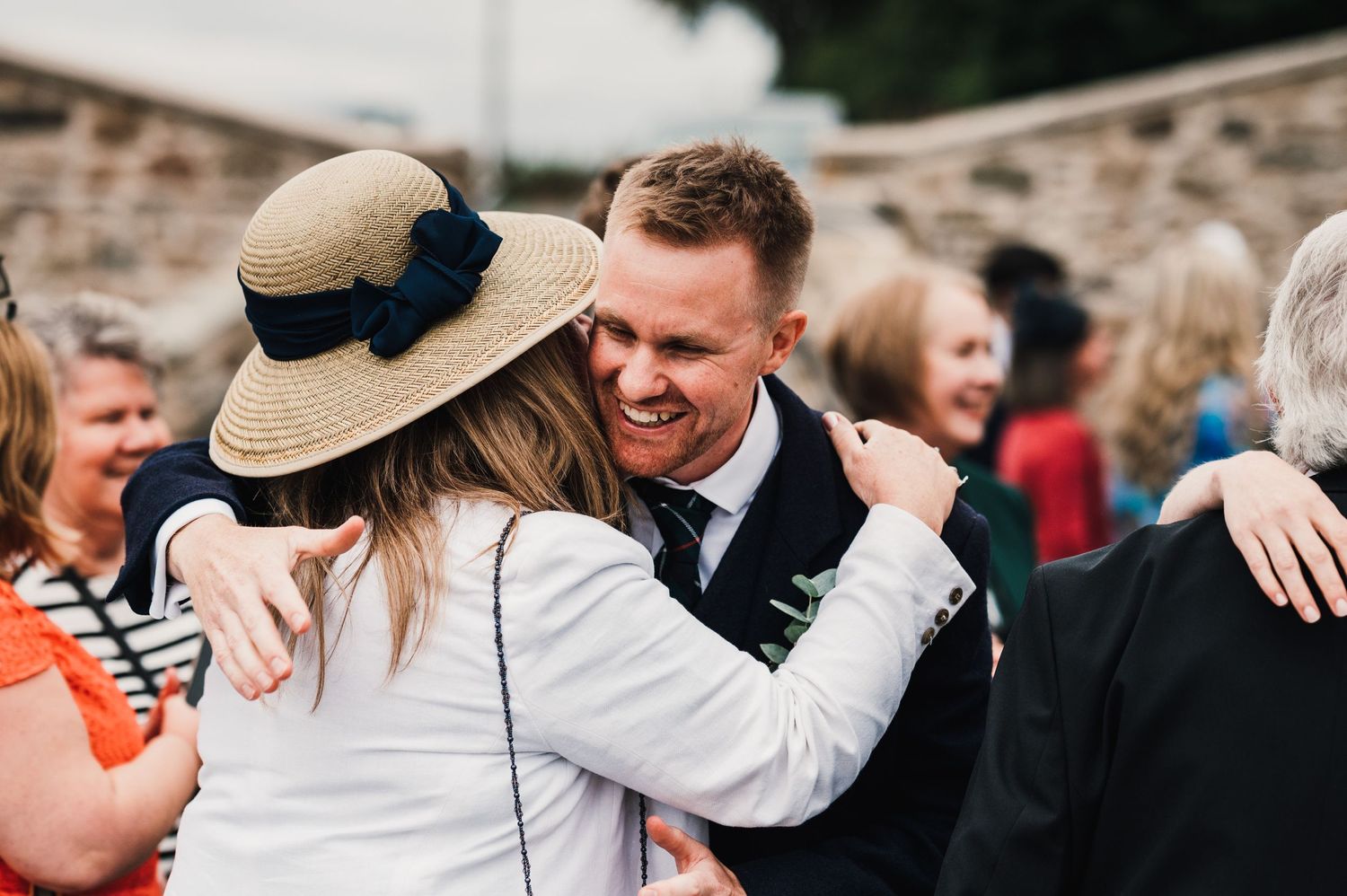 Wedding guest in straw hat embraces others during outdoor reception celebration.