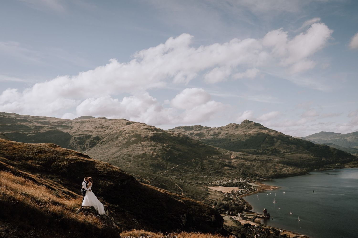 A distant wedding portrait on a hillside overlooking a scenic mountain lake.