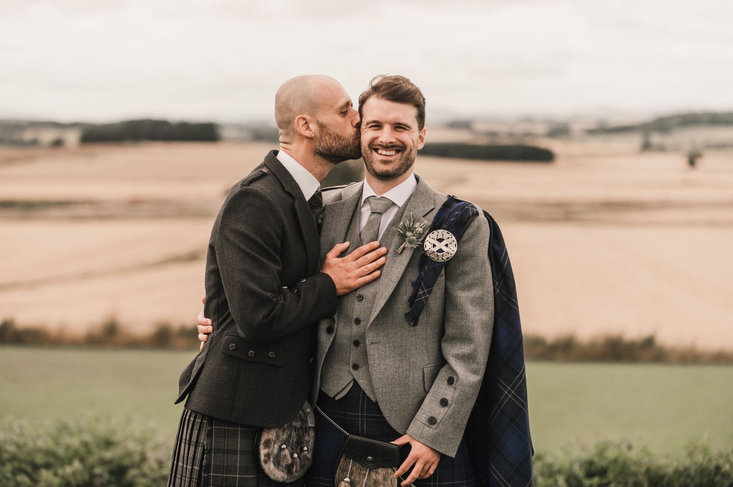 Two people in Scottish wedding attire sharing intimate moment against rural landscape.