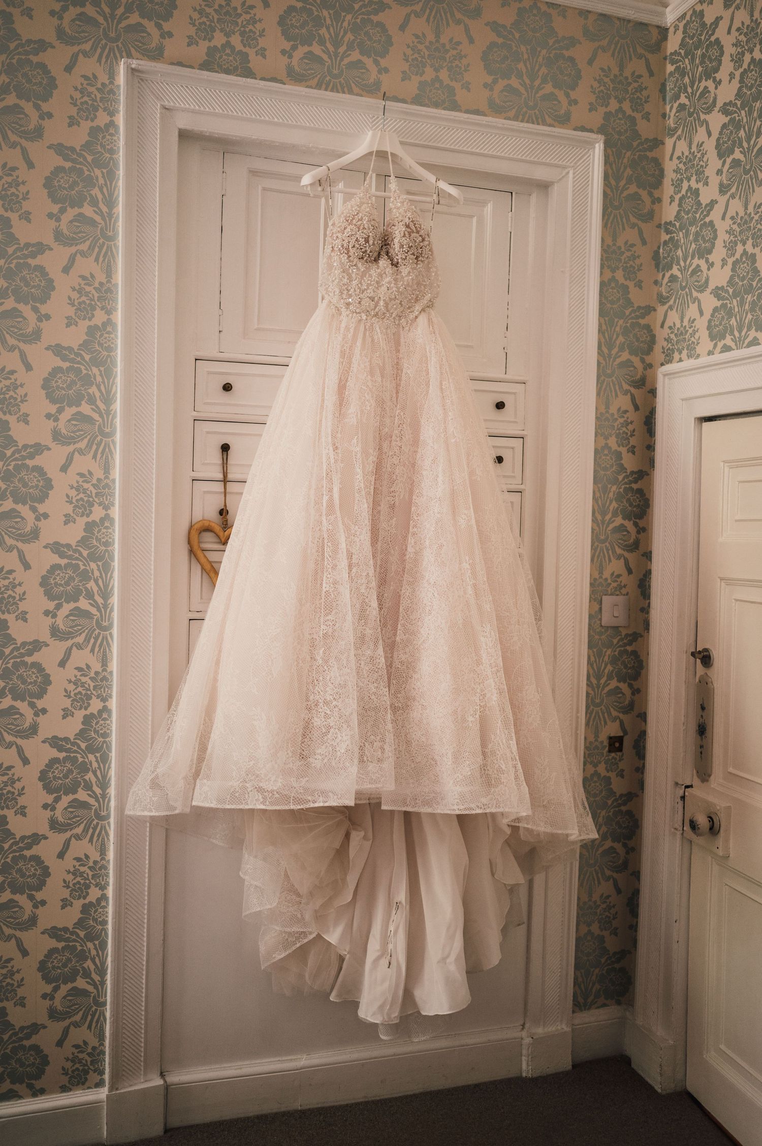 An elegant lace wedding dress with tulle skirt hangs on display against blue damask wallpaper.