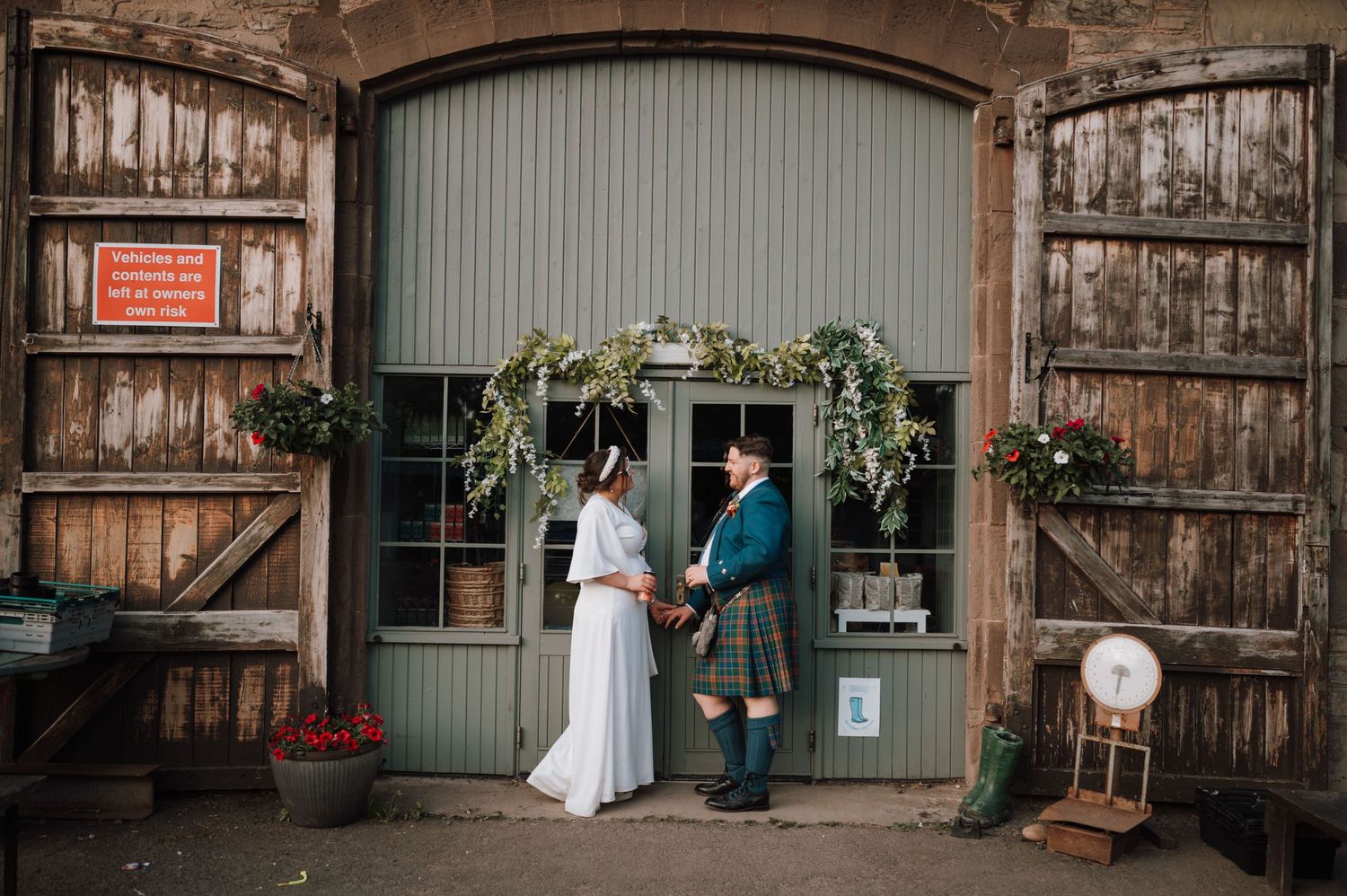 Wedding couple poses at rustic barn entrance decorated with floral arch.