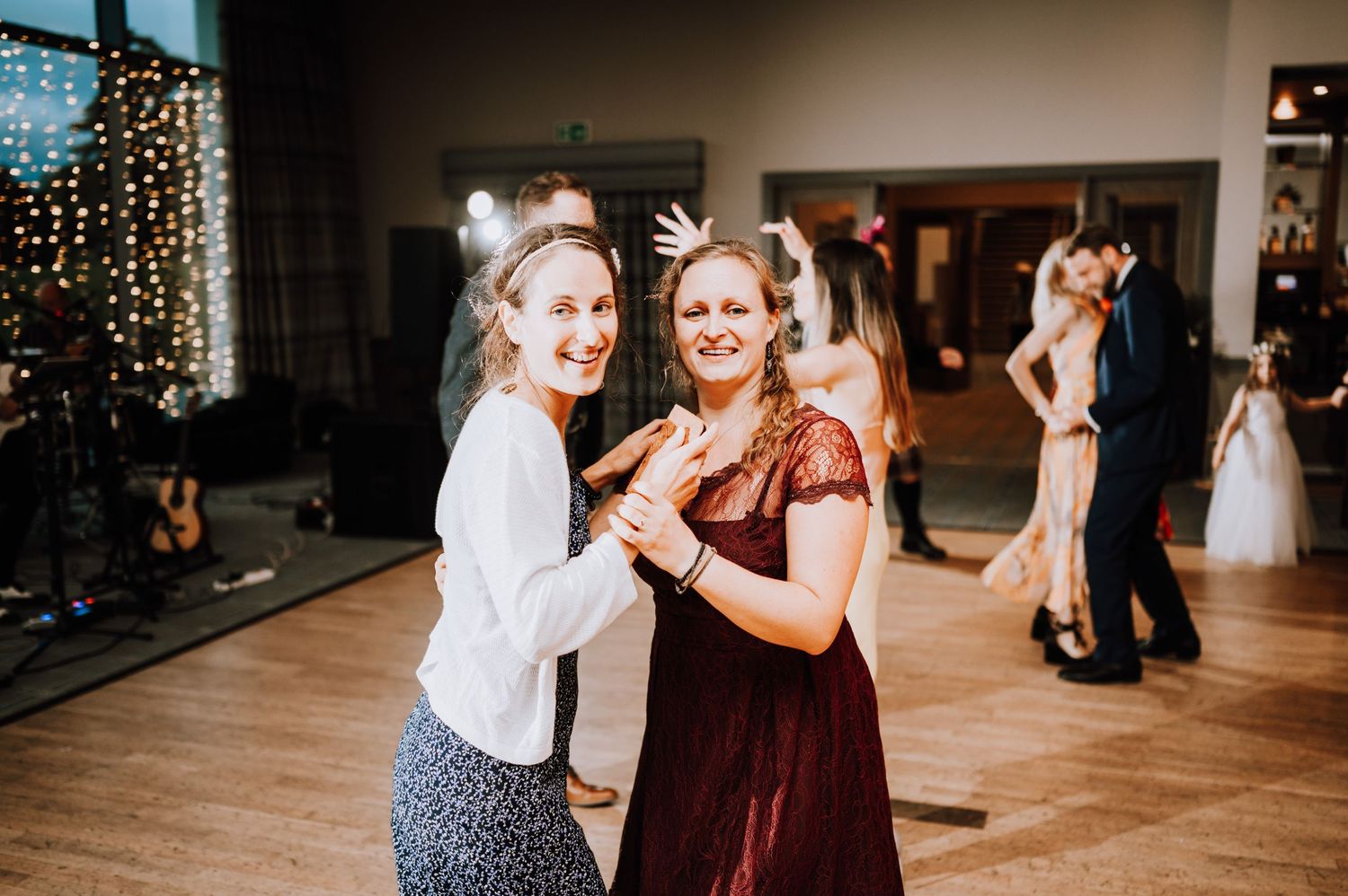 Wedding guests enjoy dancing at a reception with string lights and wooden floors.