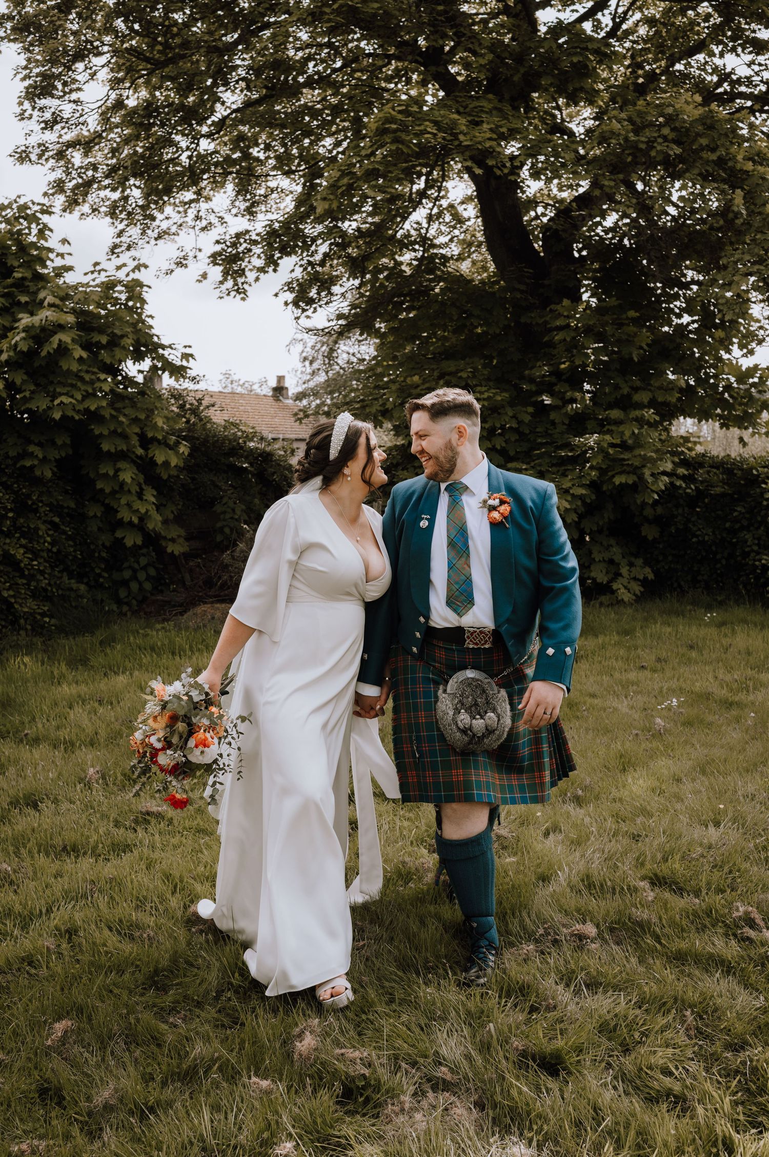 Bride in white dress and groom in blue kilt walking together in garden setting.