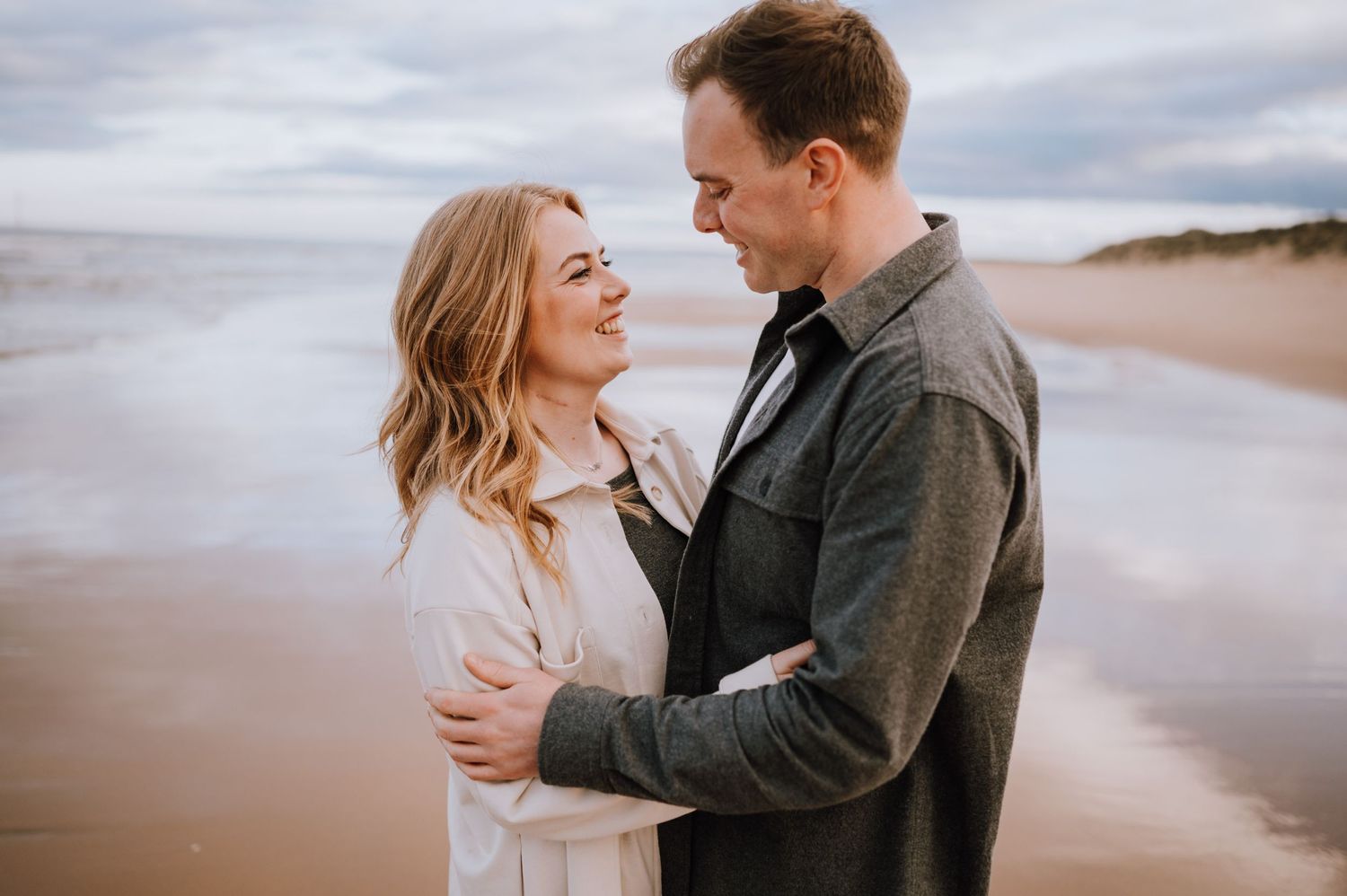 A romantic couple embraces on a cloudy beach during a coastal photoshoot.