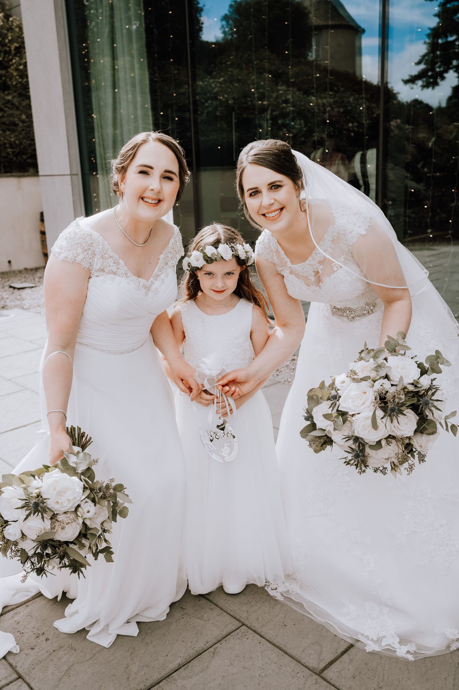Three people in white dresses pose together on a porch with wedding bouquets.