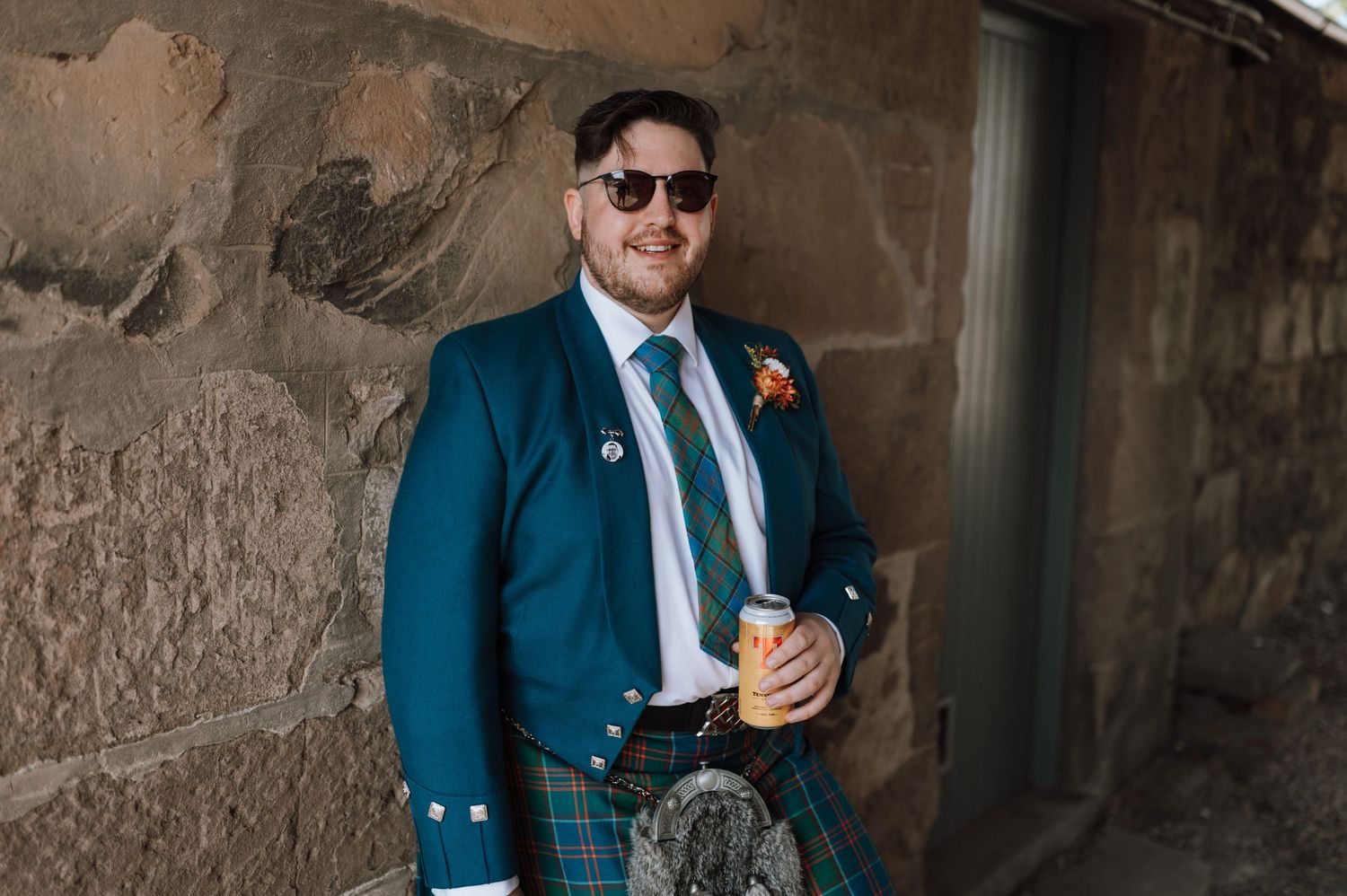 Wedding guest in traditional Scottish attire featuring teal jacket and kilt poses against stone wall.