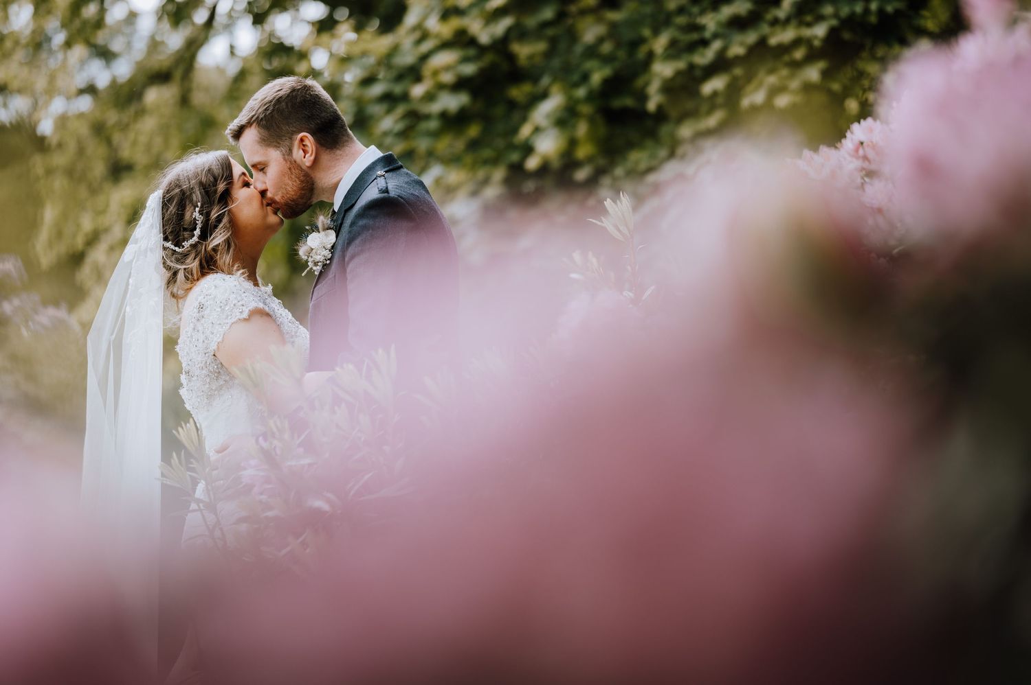 Romantic wedding kiss captured through soft pink blurred flowers in a garden setting.