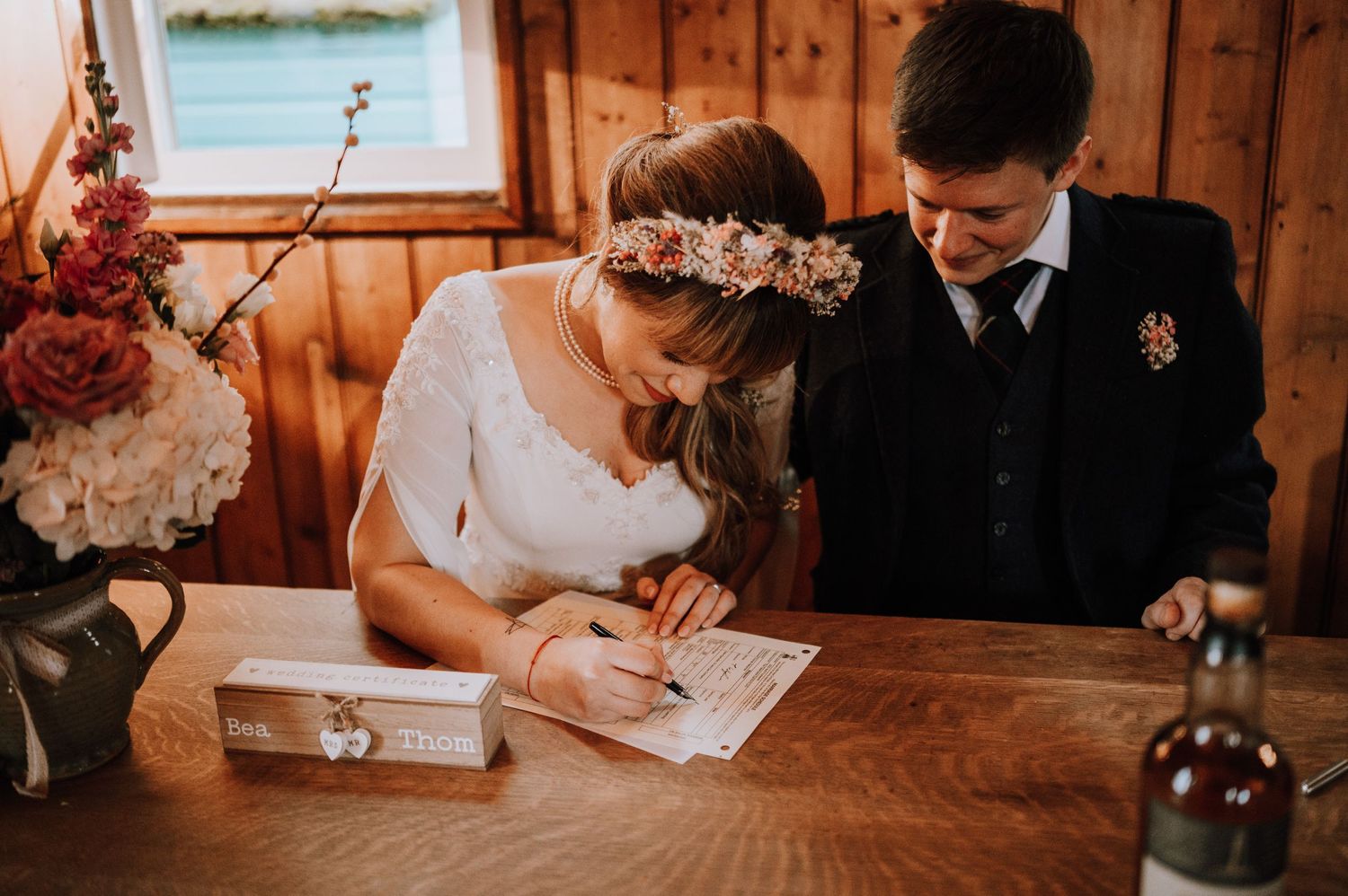 Couple signing marriage documents at rustic wooden table with floral decorations.