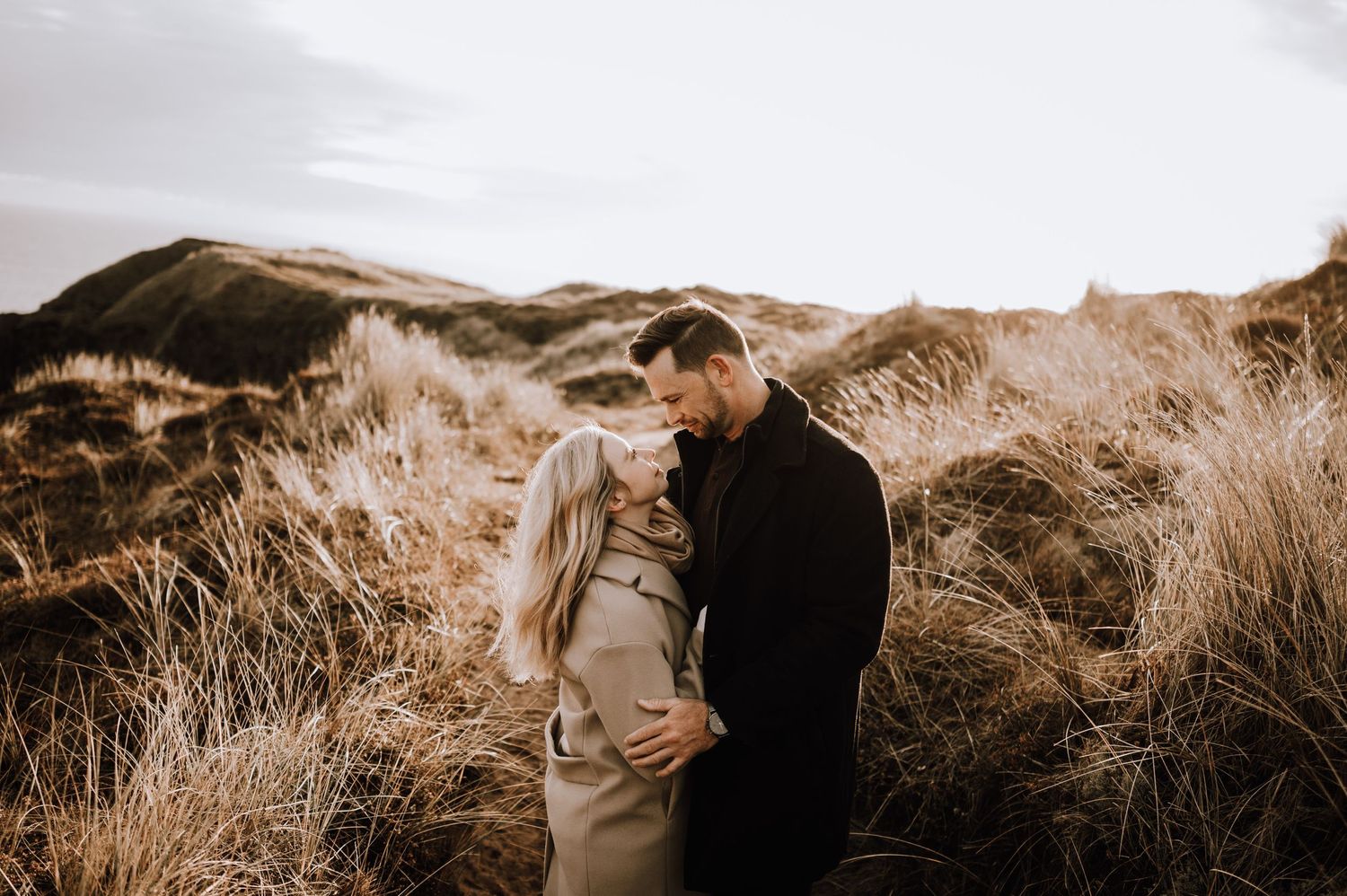 A couple shares an intimate moment on grassy coastal hills during sunset.