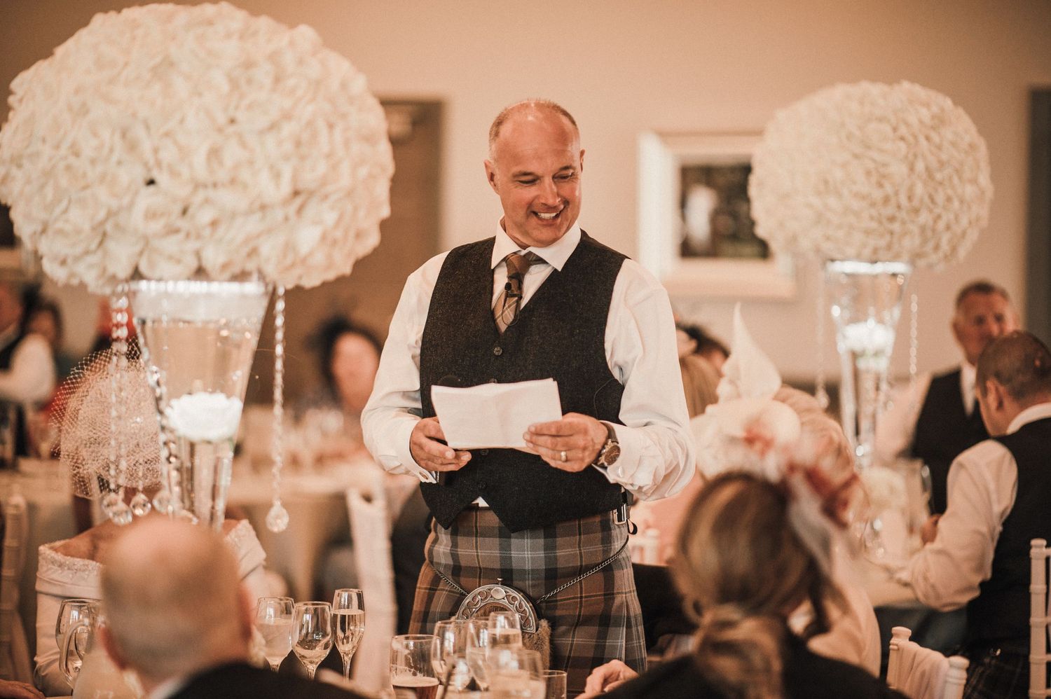 A speaker in formal attire reads from papers at an elegant reception with white floral centerpieces.