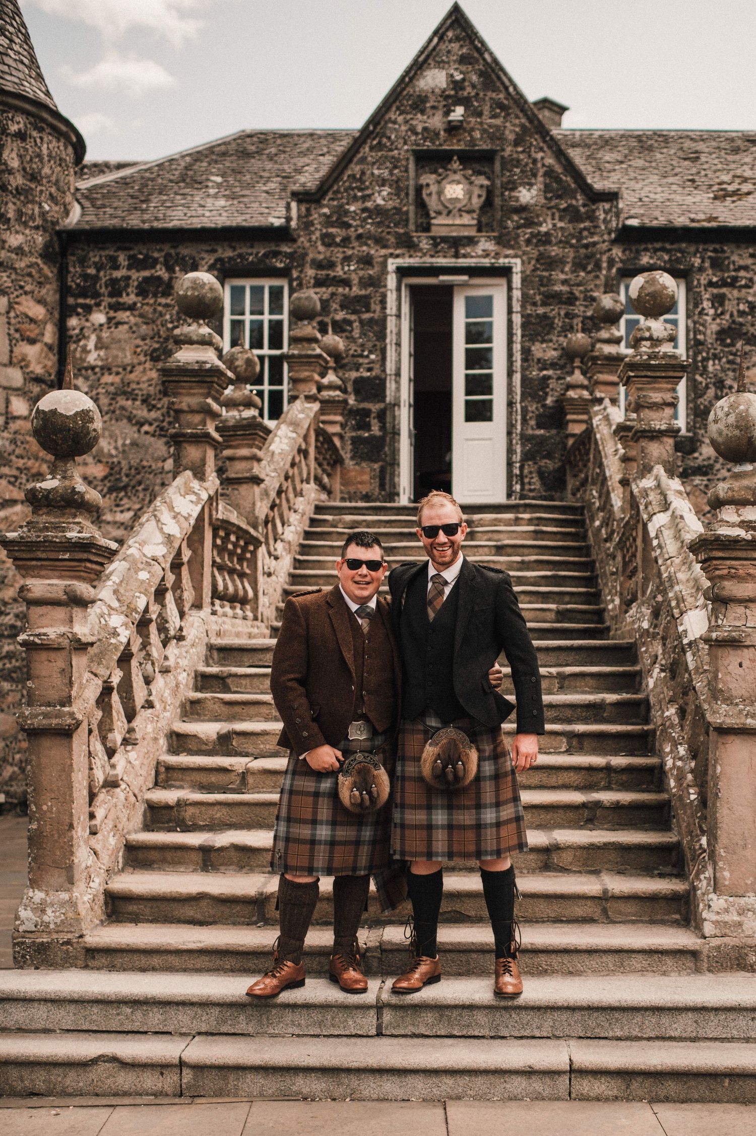Two people in traditional Scottish kilts pose on stone steps of a historic building.