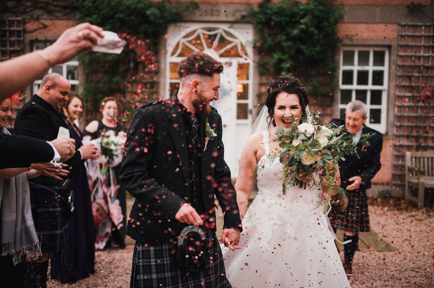 Wedding celebration outside brick building as guests throw confetti at celebrating couple.