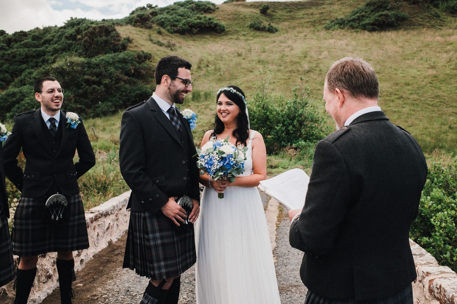 A Scottish wedding ceremony takes place on a rustic stone bridge with rolling hills in the background.