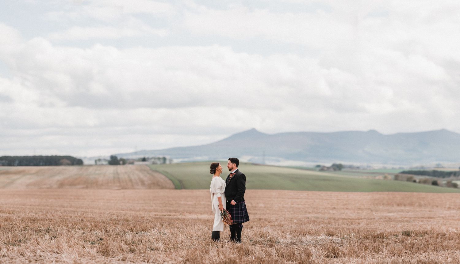 A couple stands together in a vast countryside field with mountains in the distance.