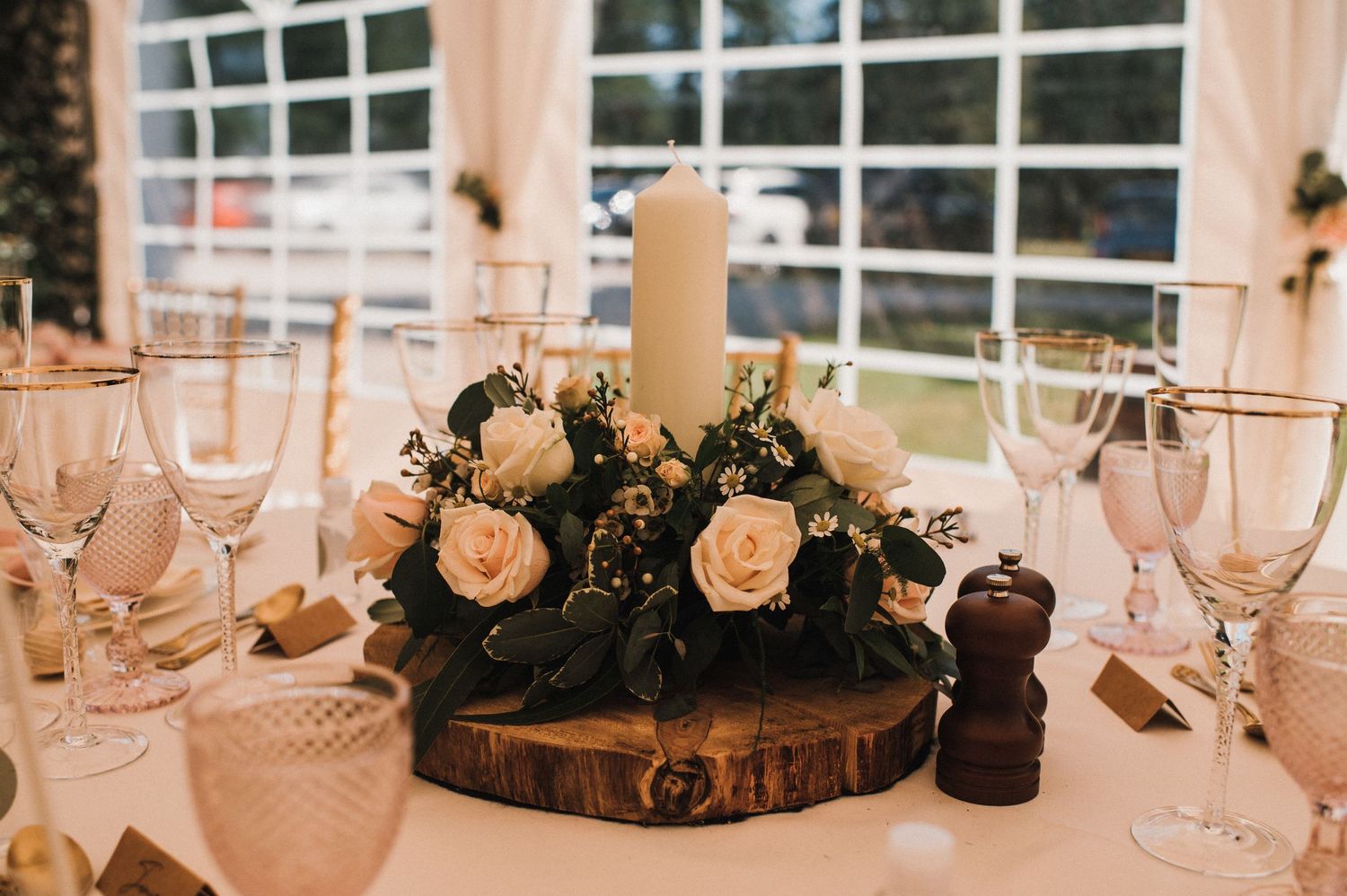 Wedding centerpiece with white roses and candle on wooden slice surrounded by glassware.