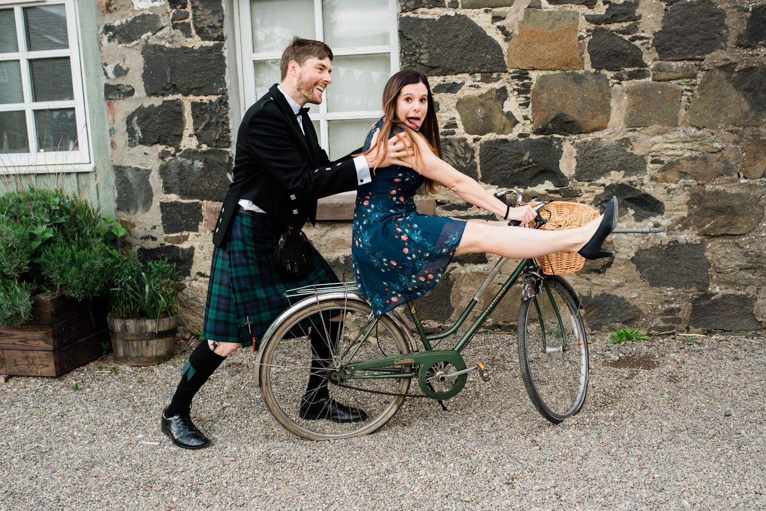 Playful wedding photo with guest in kilt pushing someone on vintage bicycle.