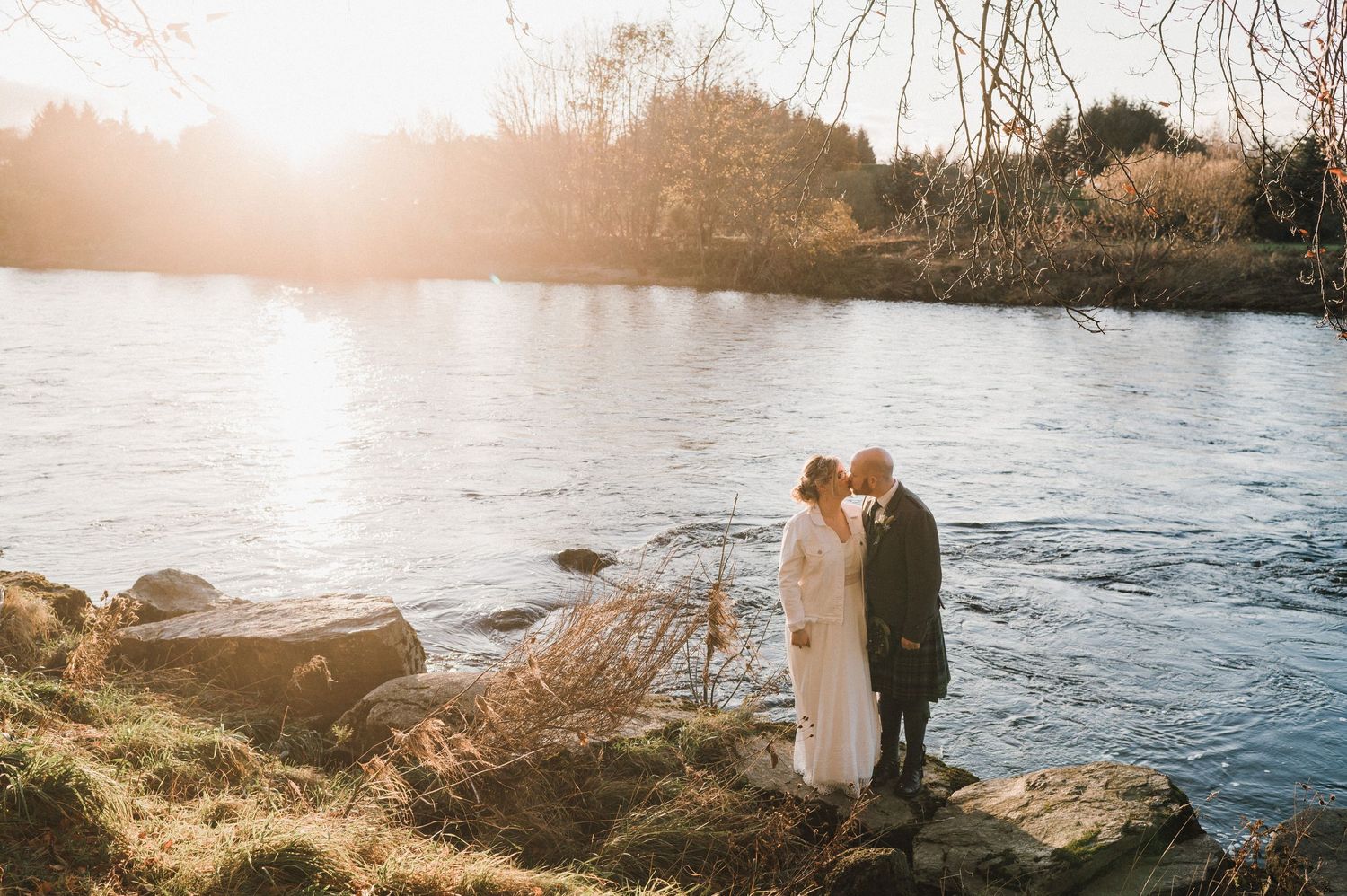 Two figures embrace on rocky riverside during golden hour with water reflections.