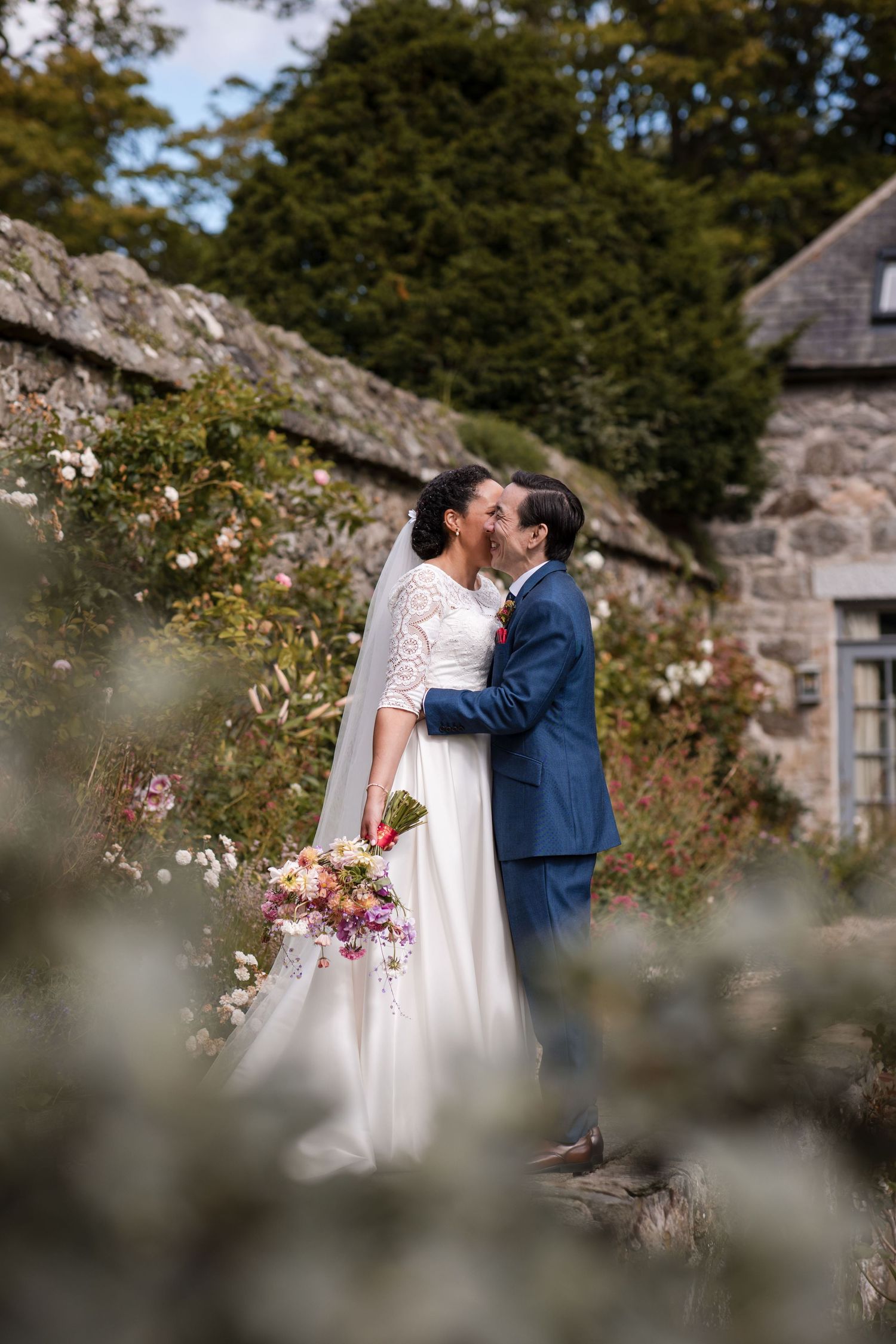 Romantic garden wedding portraits with blooming roses and stone architecture in background.