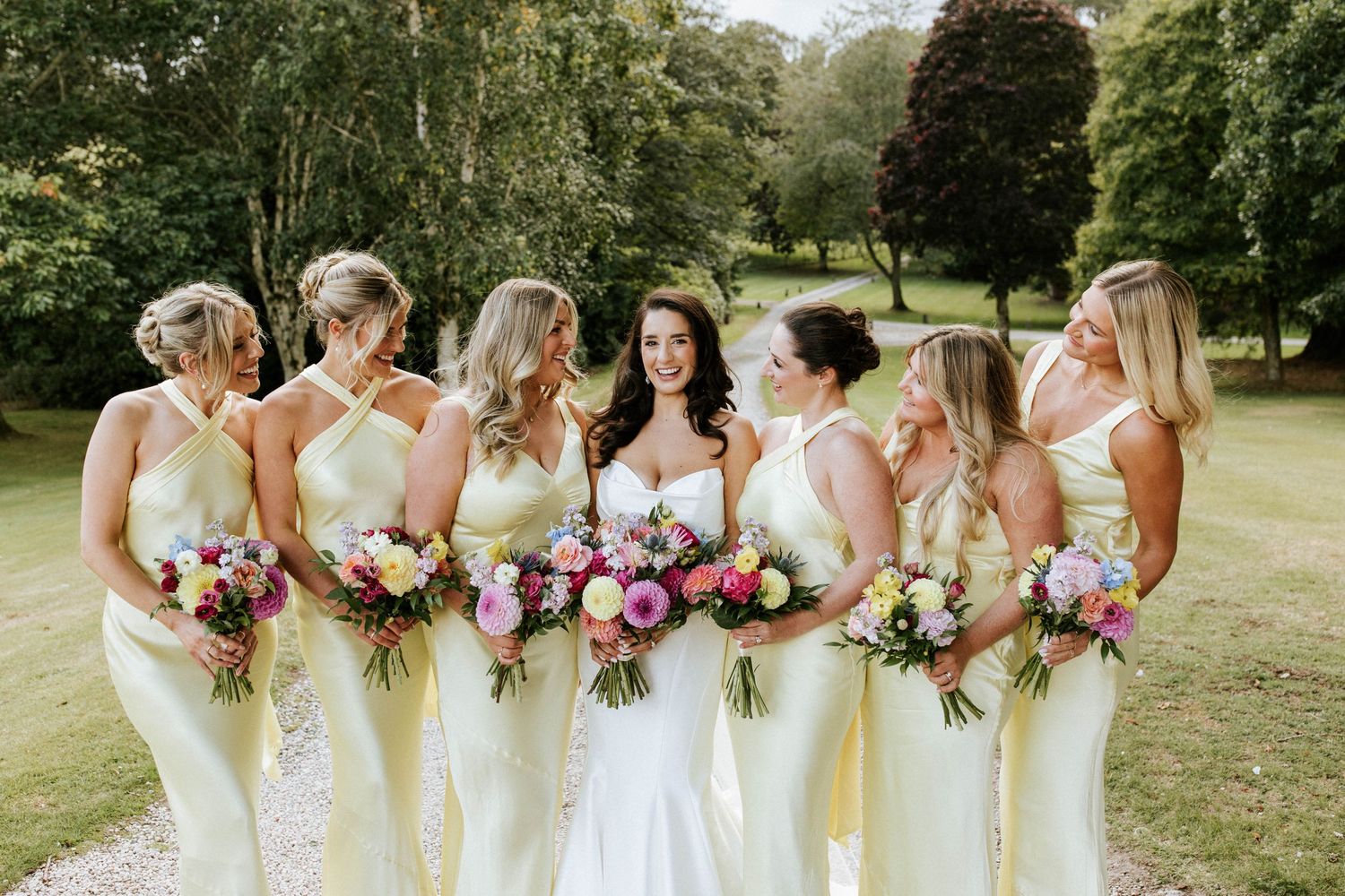 Row of bridesmaids in cream dresses holding colorful bouquets in garden setting.